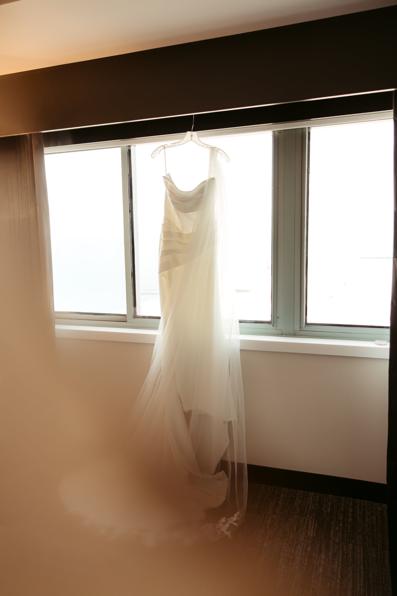A white wedding dress hanging on a hanger in front of a bright window.