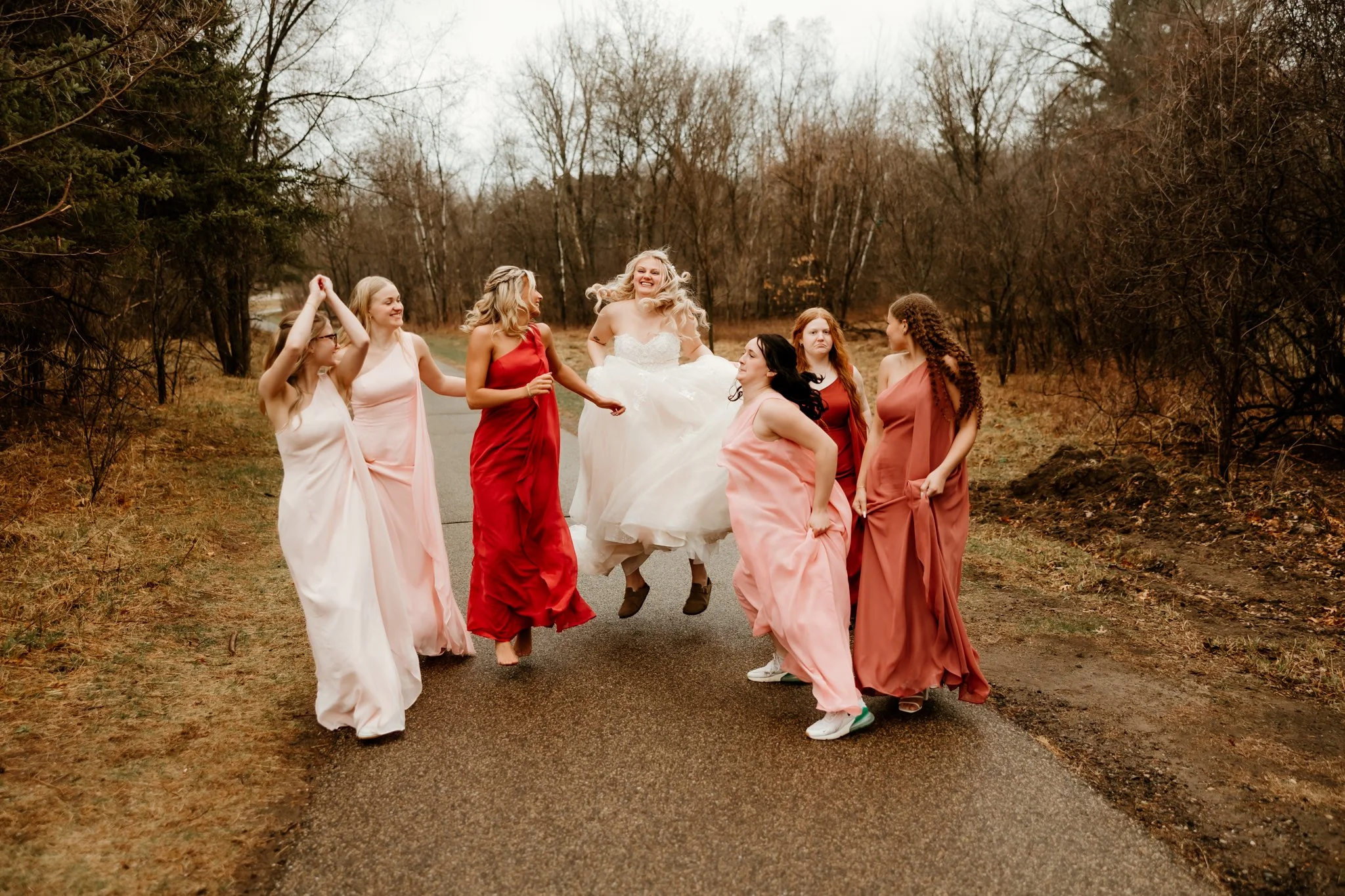 A bride in a wedding dress jumping in the air surrounded by six women in colorful dresses on a rural path with leafless trees.