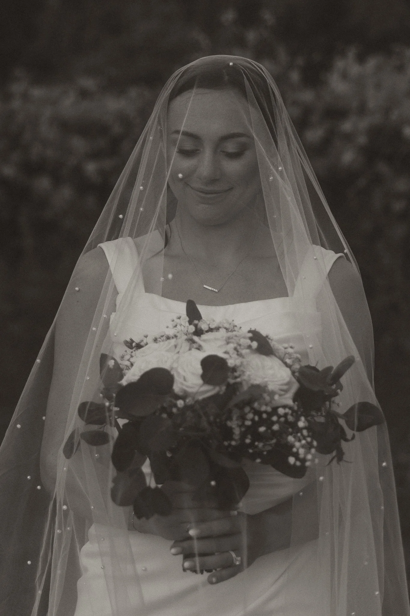 A woman in a wedding dress holding a bouquet of flowers, with a veil covering her face, smiling with closed eyes.
