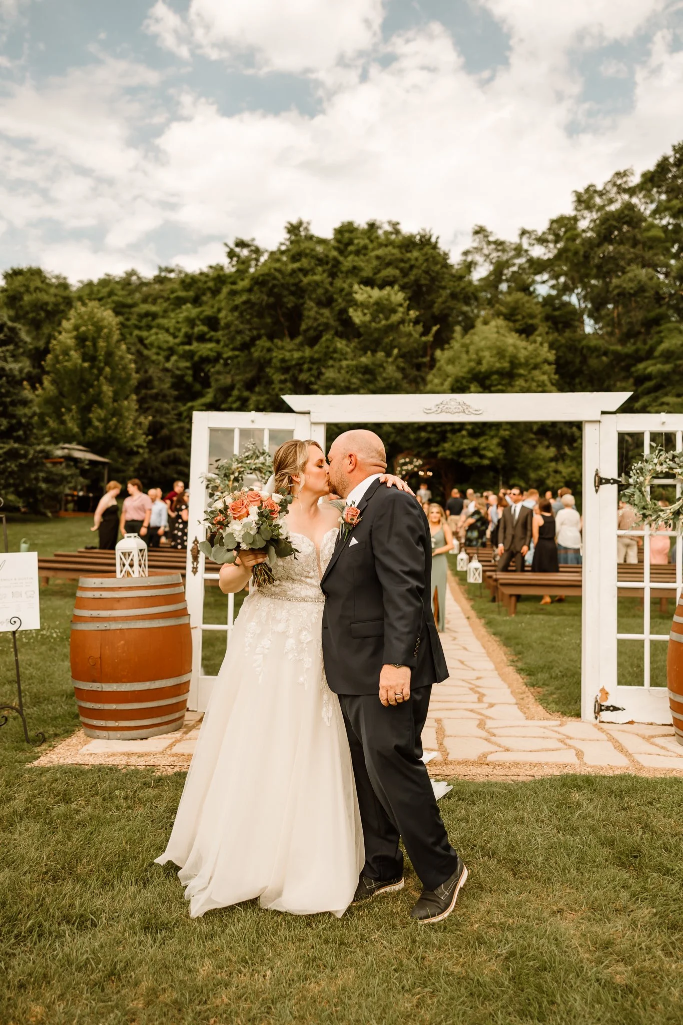 A newlywed couple kissing at their outdoor wedding ceremony with guests in the background.