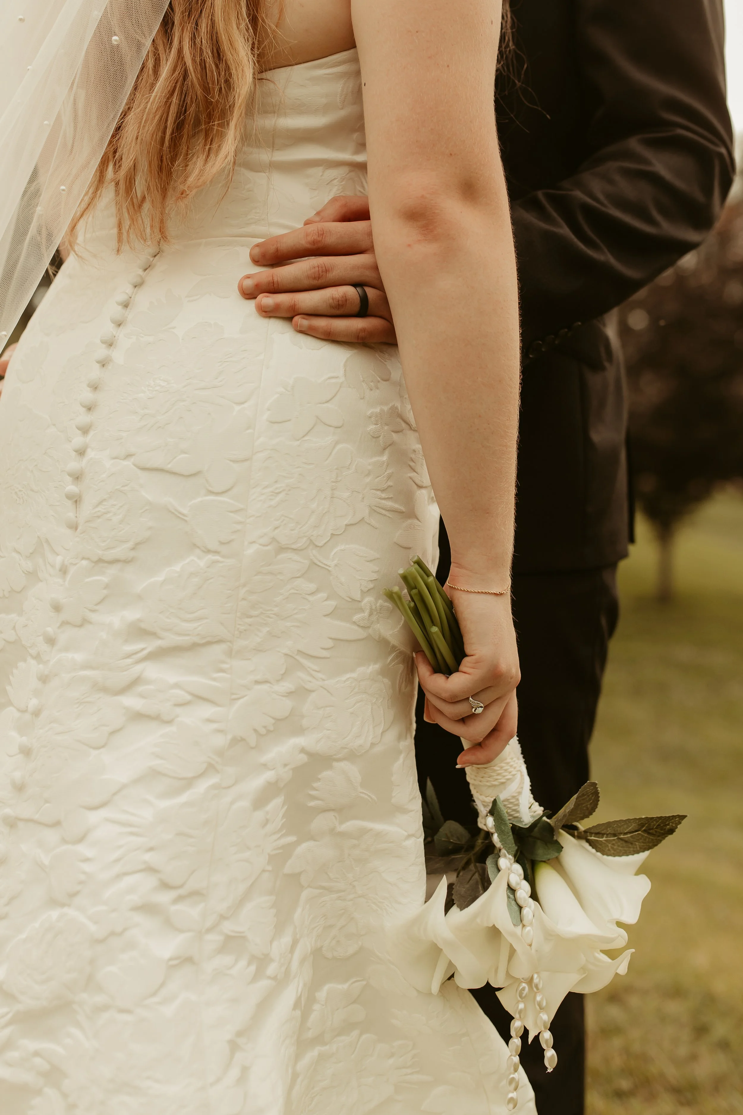 A bride holding a white flower bouquet and a groom standing behind her with his hand on her waist.