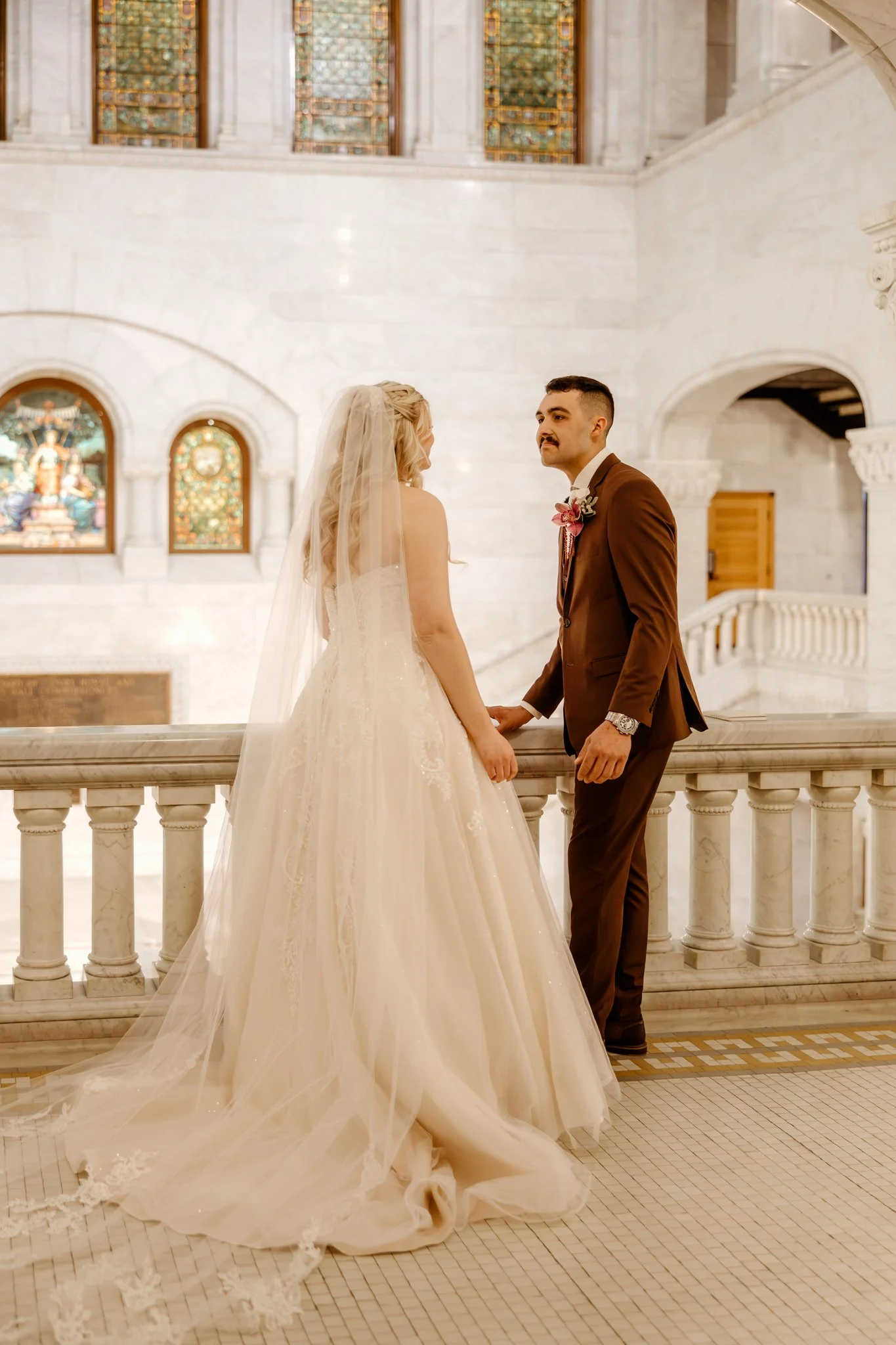 A bride and groom holding hands inside a grand church with stained glass windows, marble walls, and a staircase in the background.