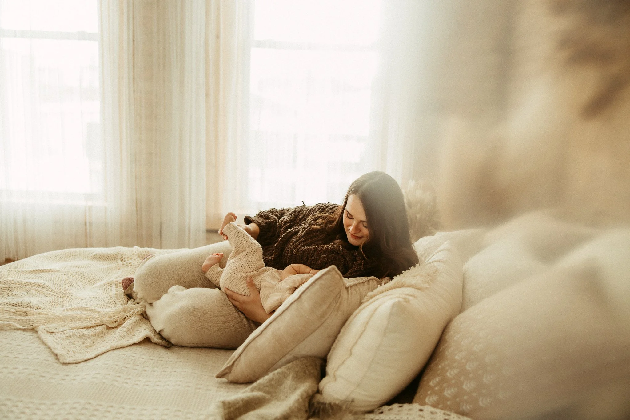 A woman lying on a bed, smiling and cradling a baby, in a cozy room with natural light and cream-colored furniture.