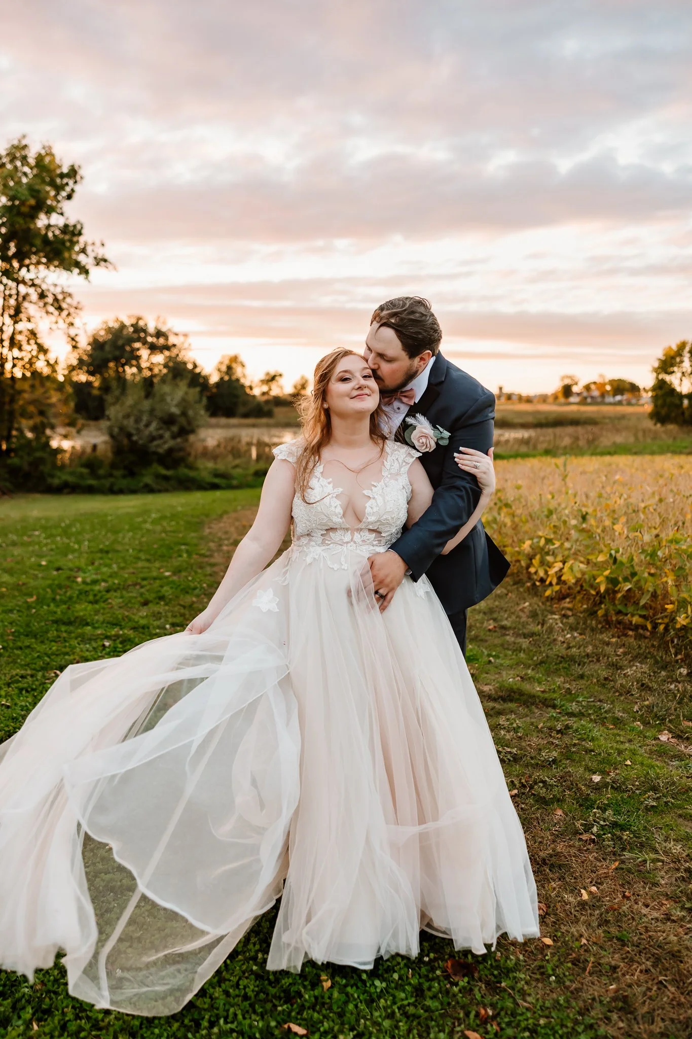 A newlywed couple standing outdoors at sunset; the bride wears a flowing wedding gown and the groom is in a dark suit, sharing a tender moment in a rural setting.