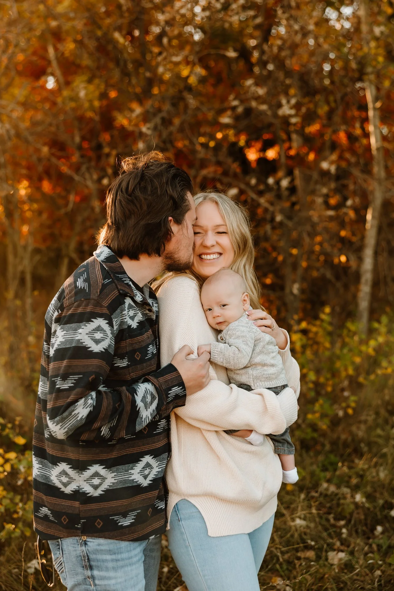 A happy family enjoying fall outdoors, with a man kissing a woman on the cheek, a woman holding a baby girl, all smiling with autumn leaves in the background.