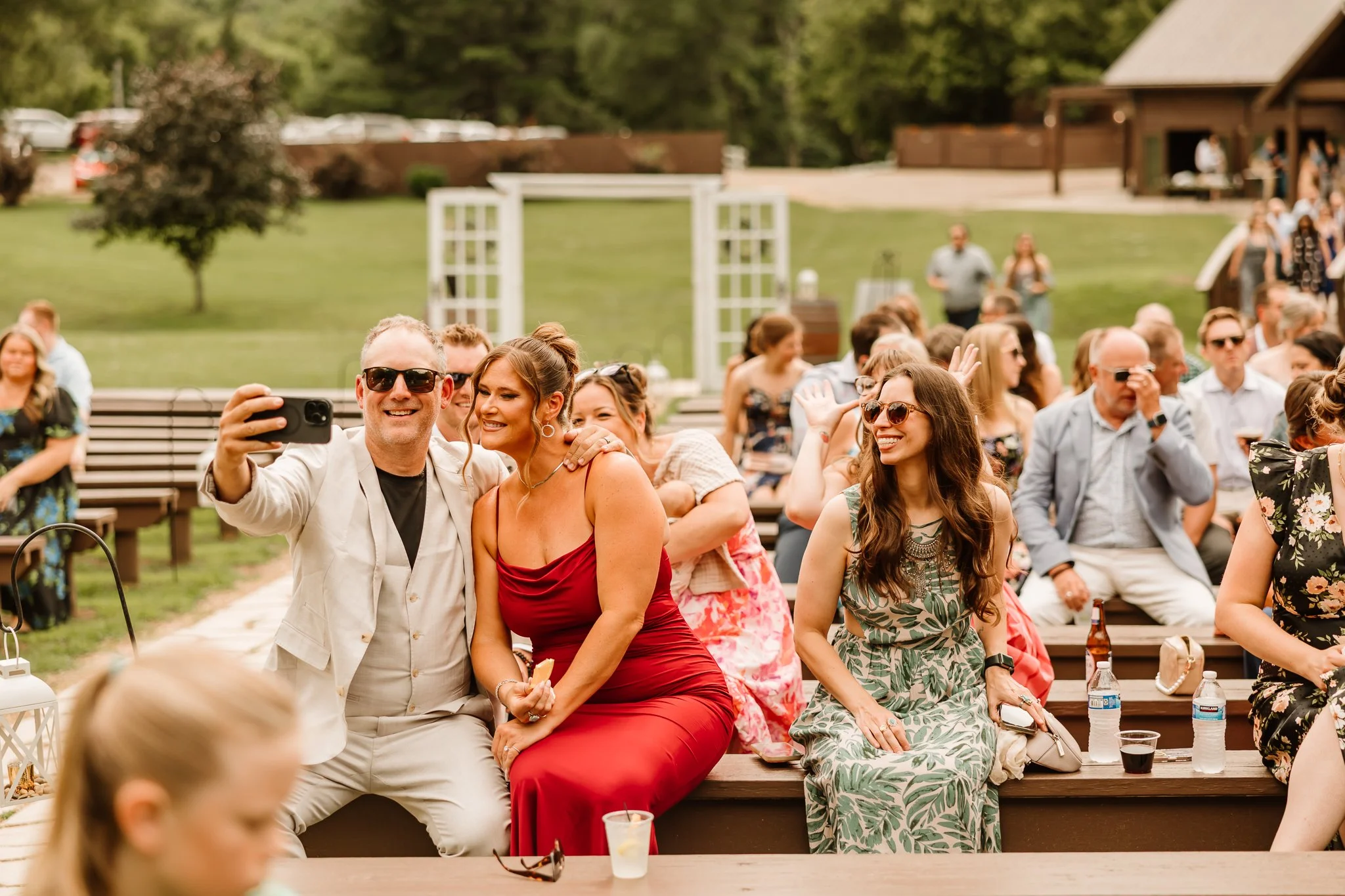 People at an outdoor wedding reception, sitting on benches, smiling, taking photos, and socializing.