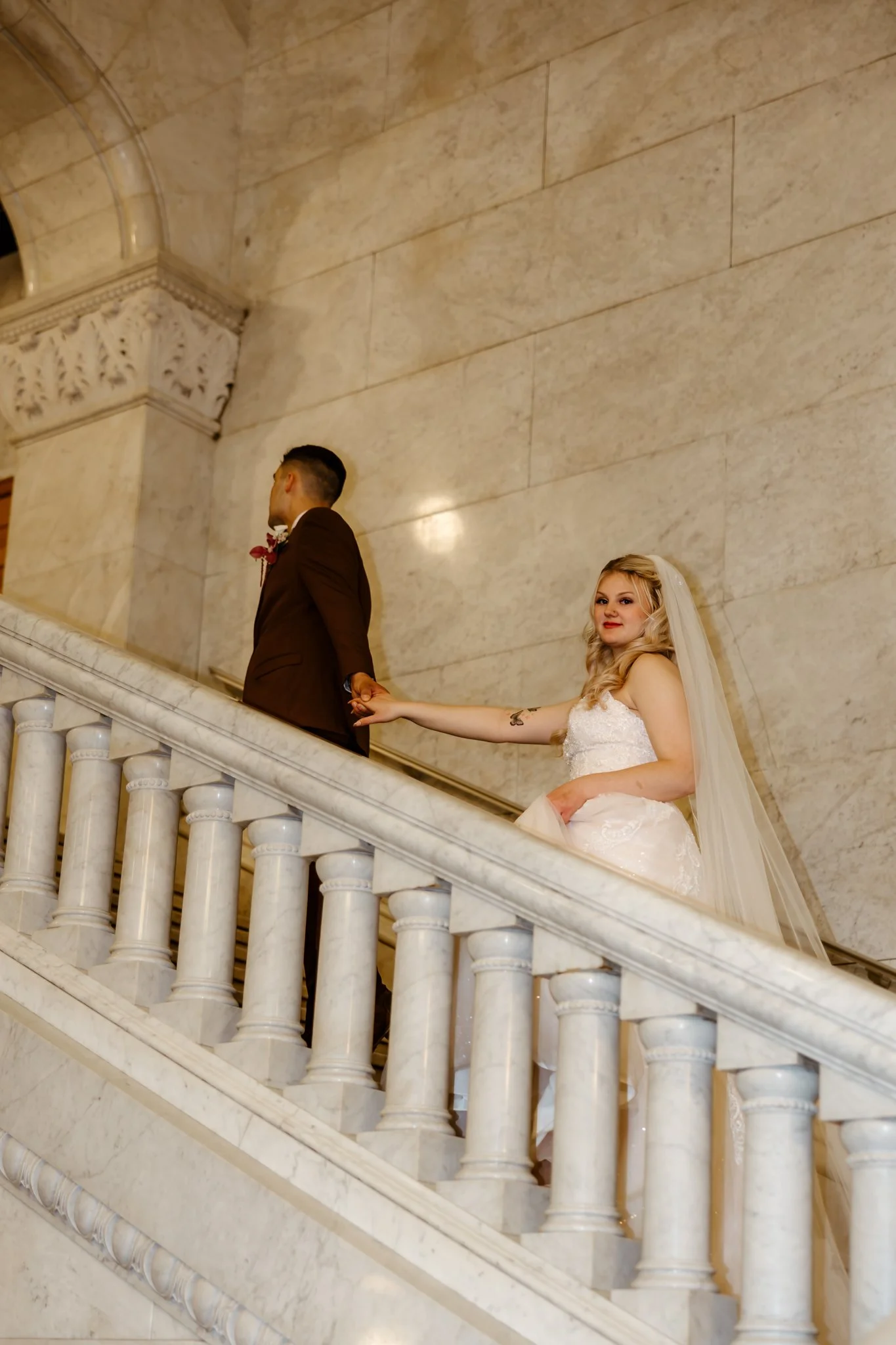 Bride and groom holding hands on a marble staircase inside a building with ornate architecture, bride in a white gown and veil, groom in a brown suit.