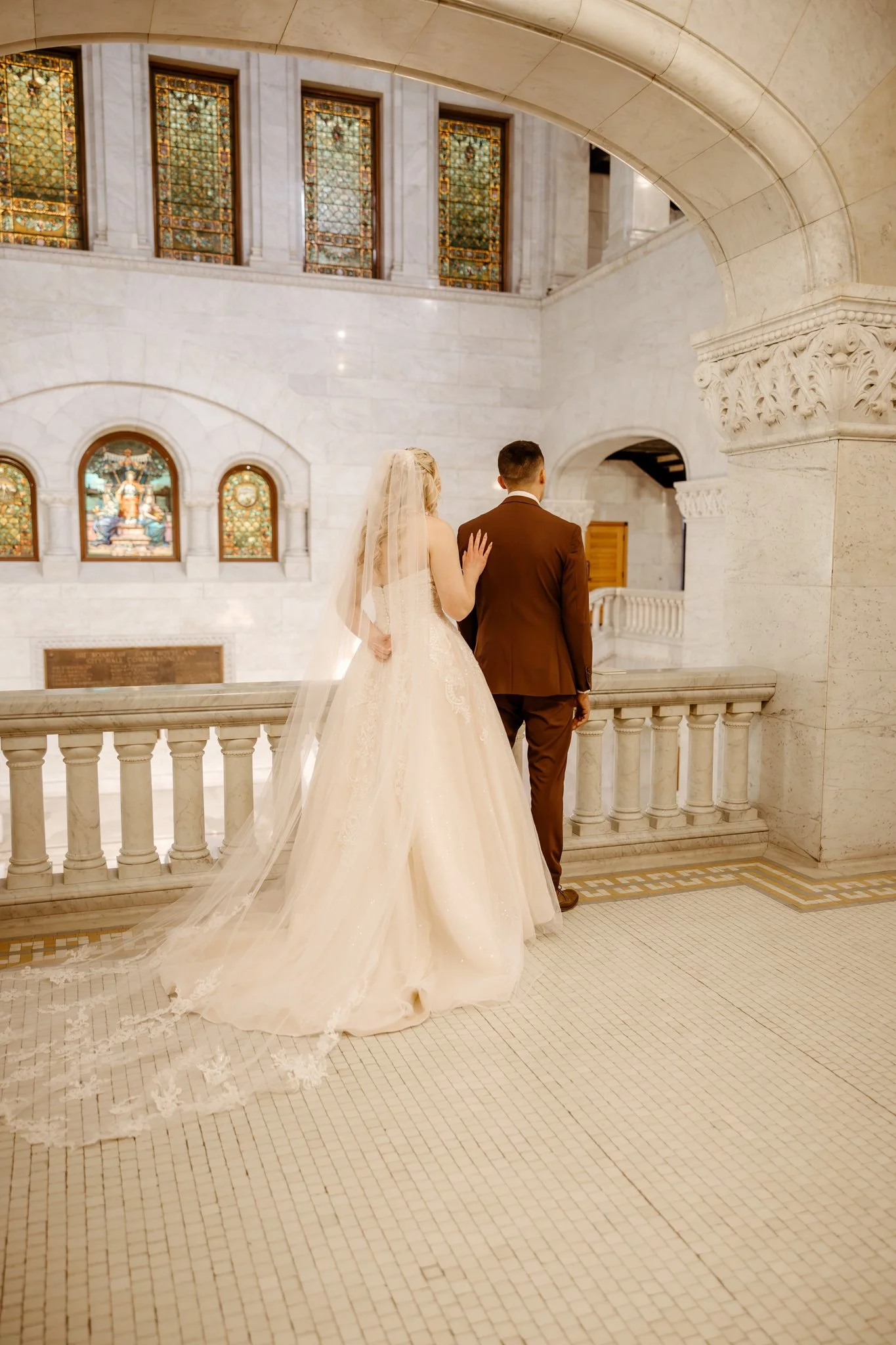 Bride and groom standing in a church, facing away, with stained glass windows and marble architecture