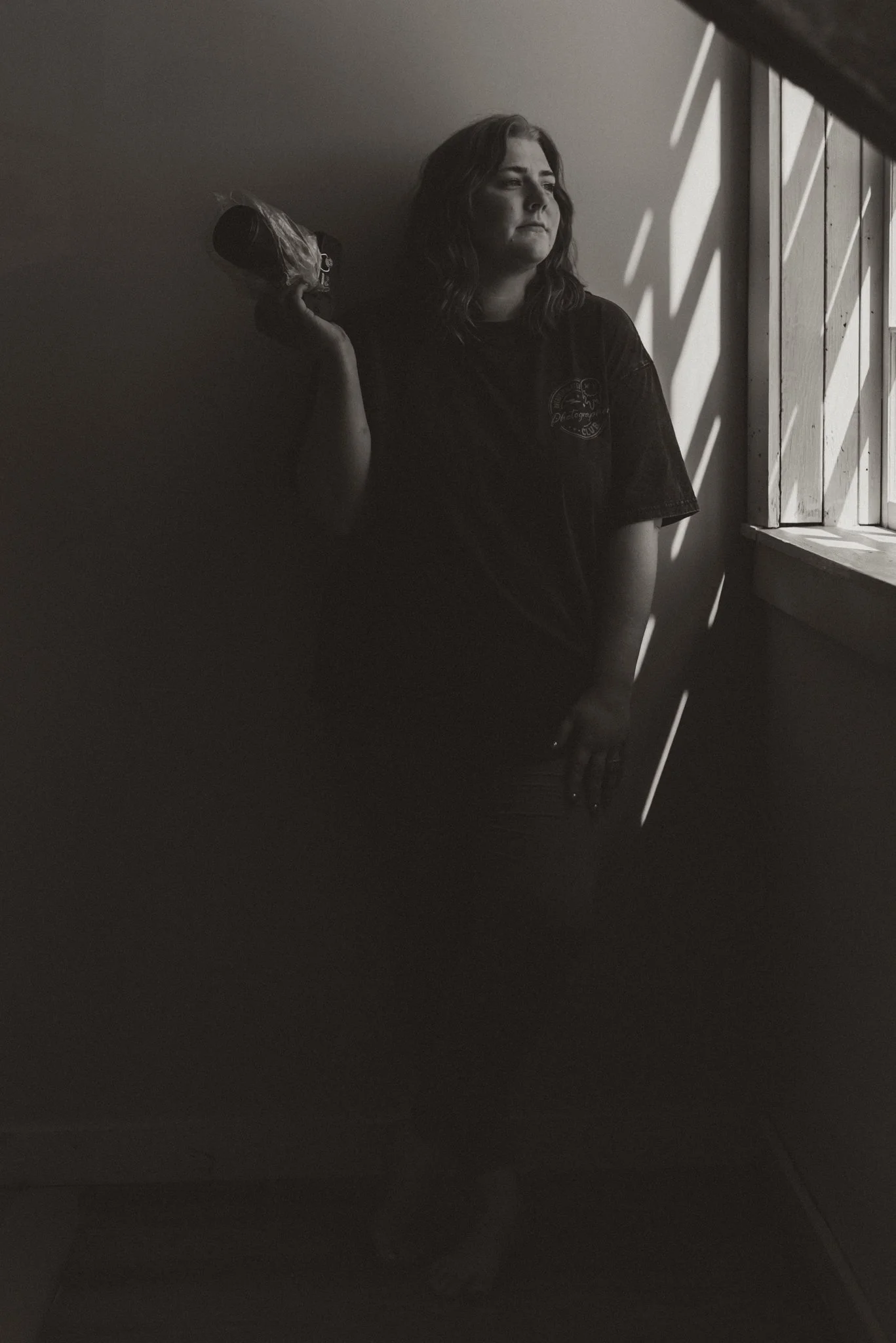 A woman standing by a window with wooden blinds, holding a canister on her shoulder, looking thoughtfully outside, with light and shadow creating patterns on the wall.