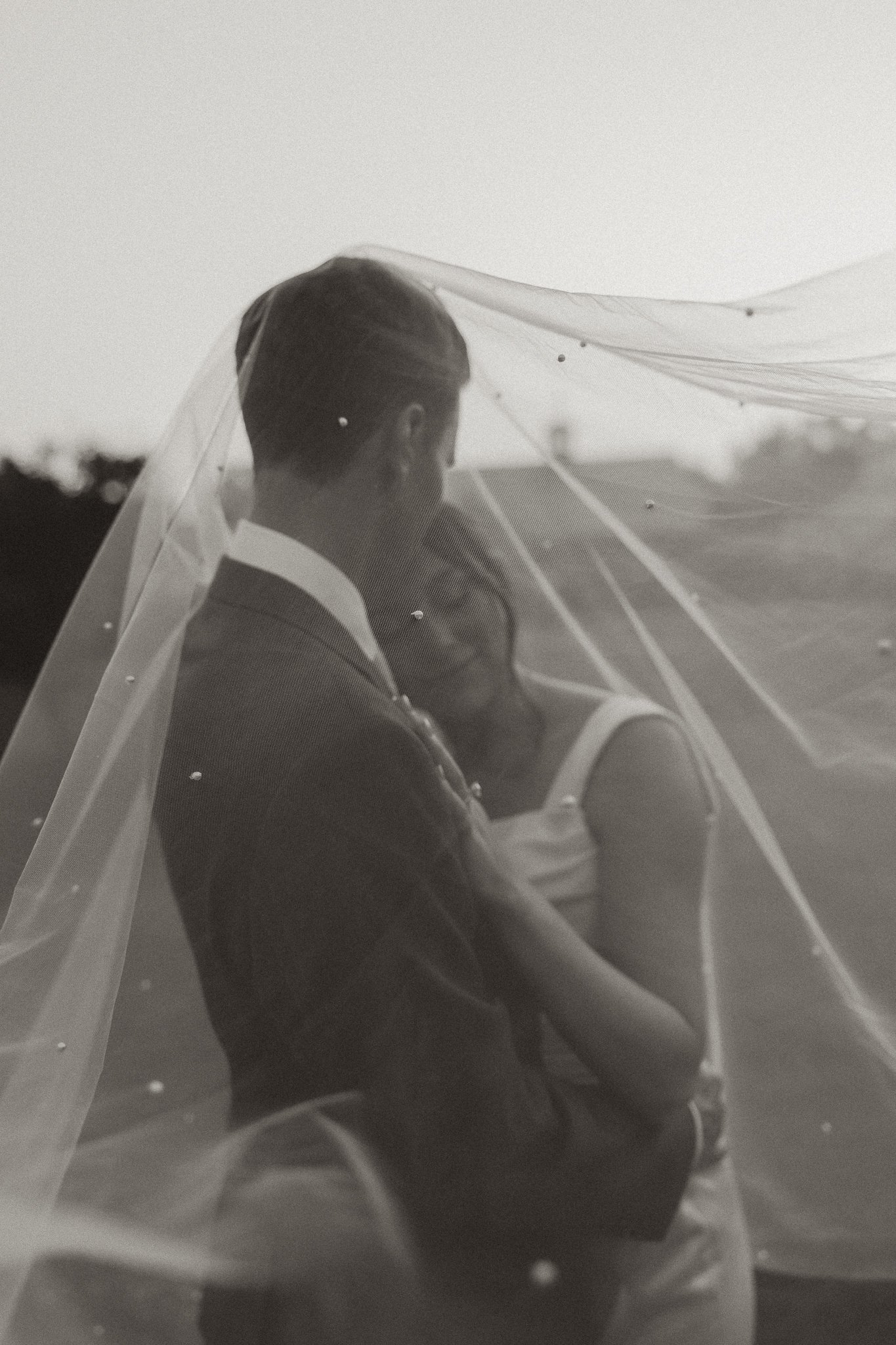 A groom holding a bride under a veil outdoors in black and white.