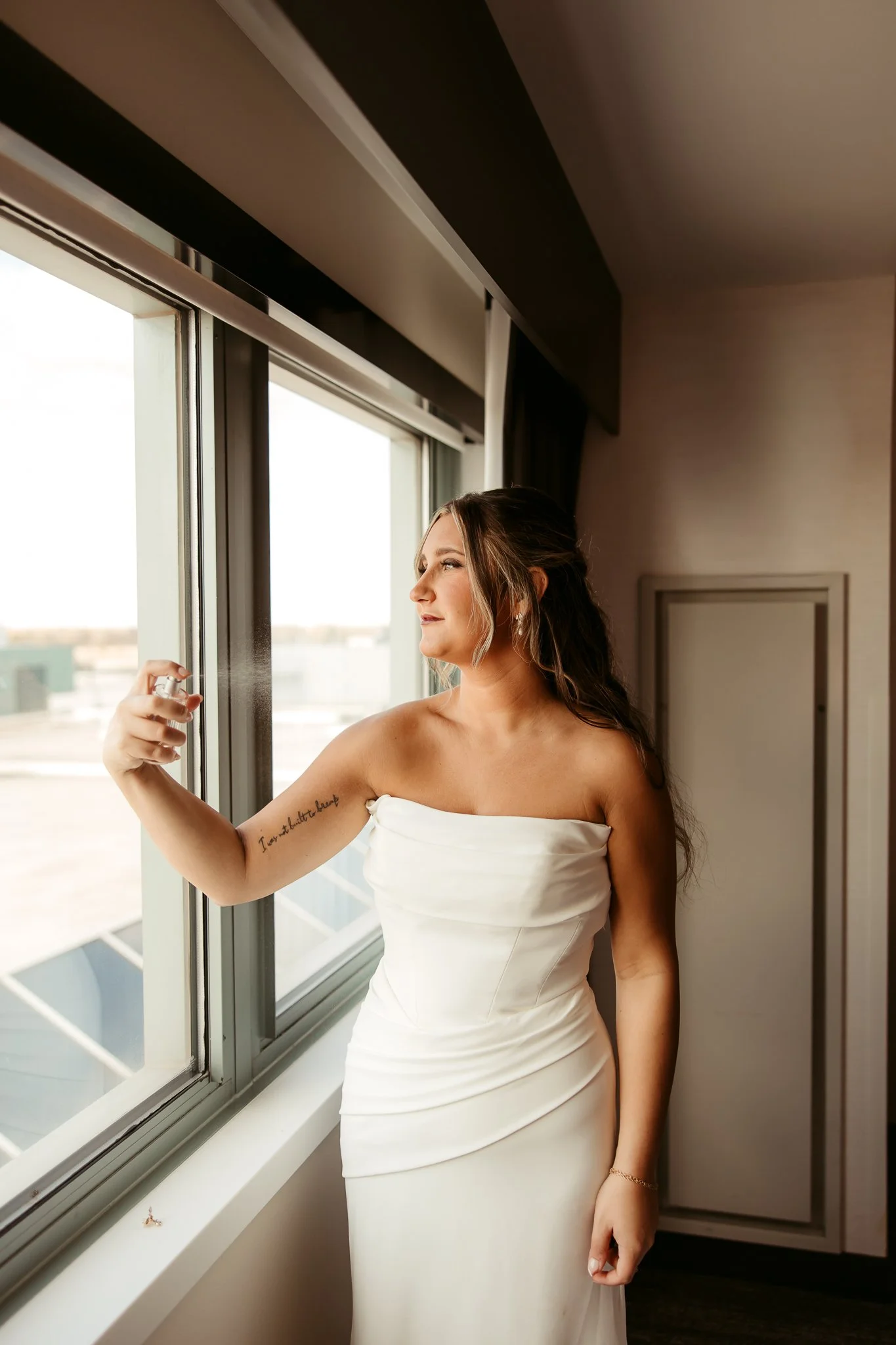 A bride in a strapless white wedding dress standing by a large window, looking outside with a serene expression.