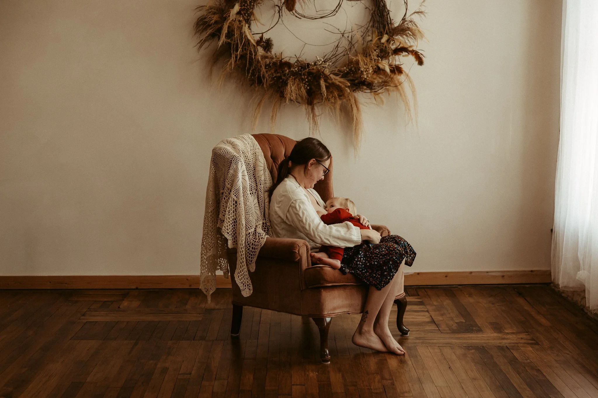 A woman with glasses sitting on a vintage armchair, holding and looking at a baby. The woman is wearing a cream sweater and a floral skirt, and the baby is dressed in red. There is a beige crocheted blanket draped over the armchair, and a decorative 