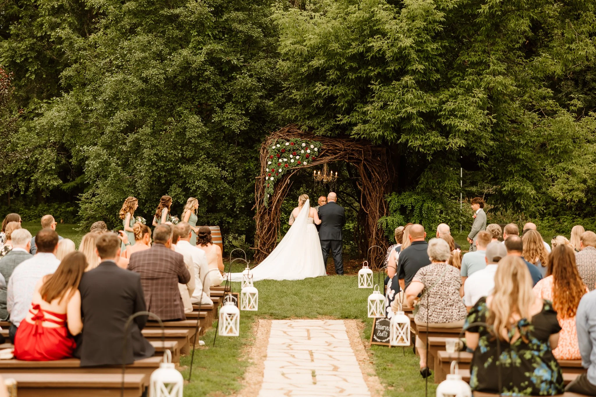 A wedding ceremony outdoors with the bride and groom at the altar, guests seated on either side, and a backdrop of lush green trees. The altar is decorated with flowers and a chandelier, and there are lanterns along the aisle.