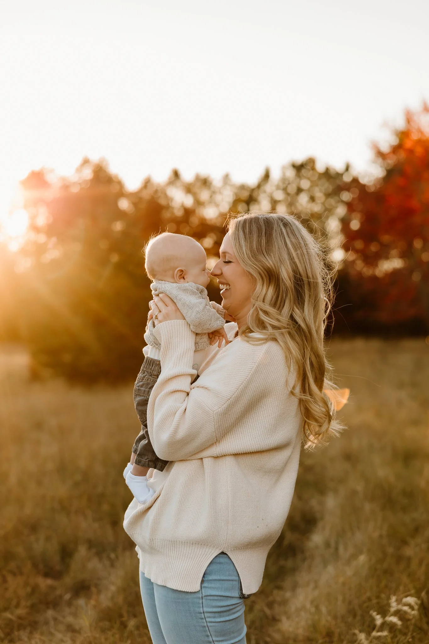 A woman holding a baby outside in a field during sunset, with autumn trees in the background.