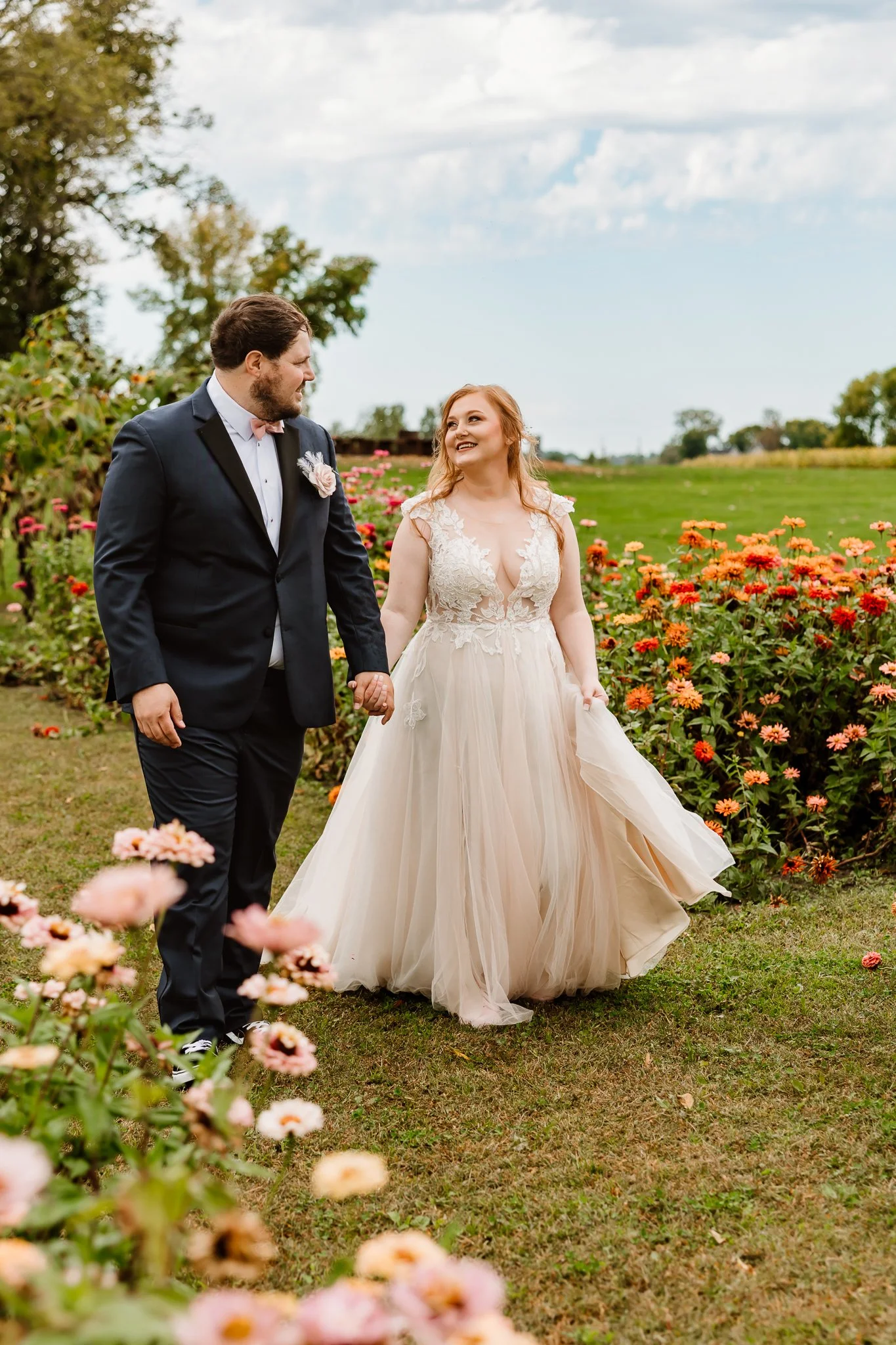 A newlywed couple holding hands and walking through a flower garden, with the bride in a white lace gown and the groom in a dark suit, under a partly cloudy sky.