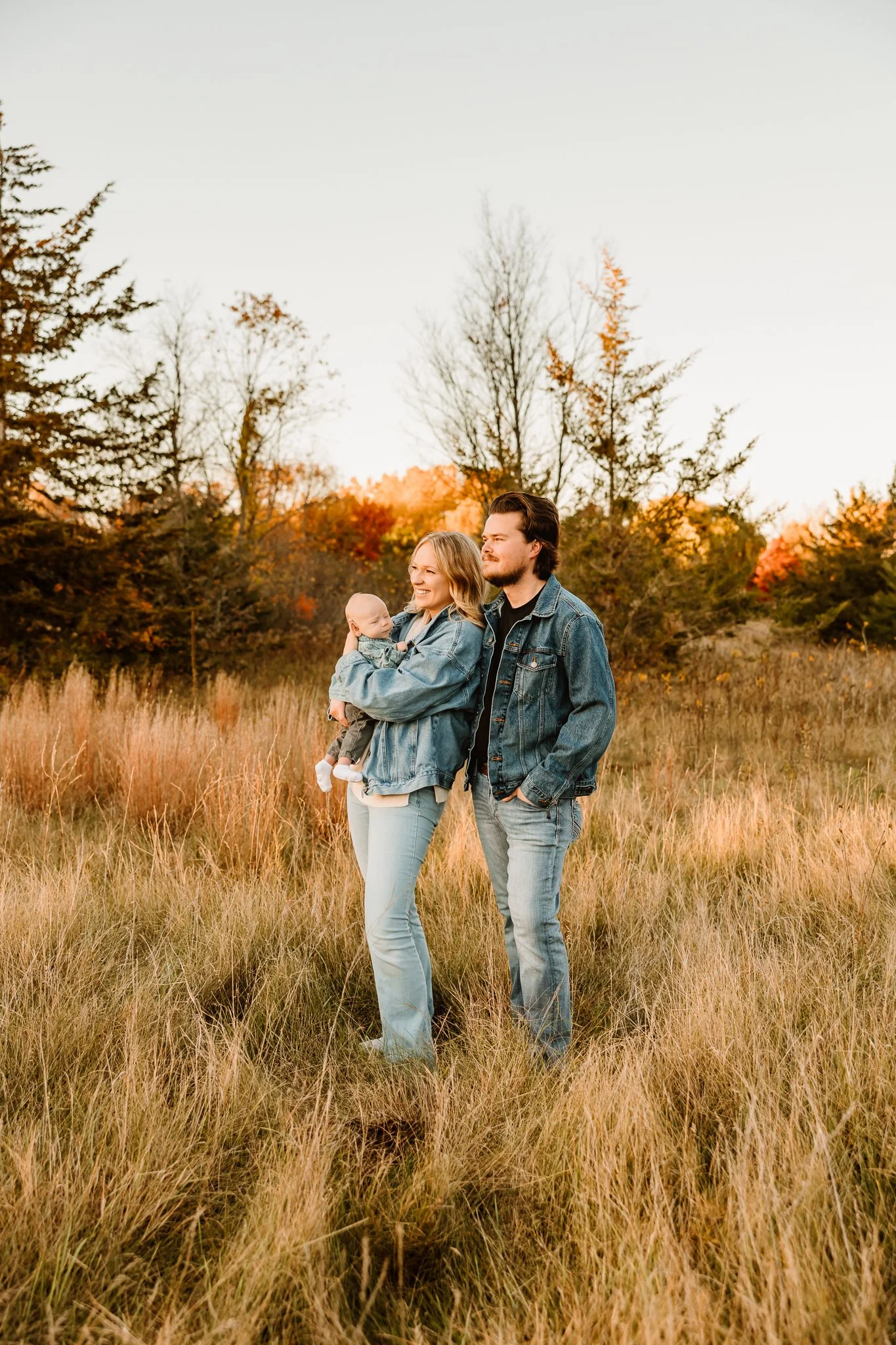 Family of three, parents and baby, standing outdoors in a grassy field during autumn, with trees with orange and red leaves in the background, all wearing denim jackets.
