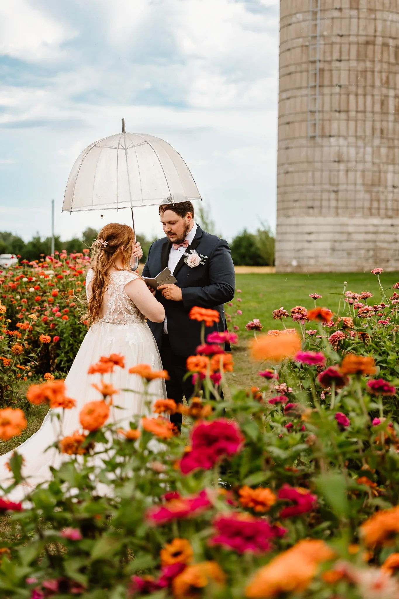 A couple getting married outdoors, with a woman in a lace wedding dress and a man in a dark suit holding a paper, sharing vows under a transparent umbrella amidst colorful flowers and a large silo in the background.