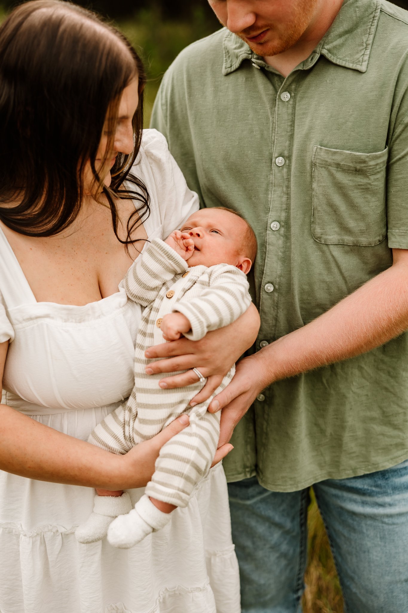 A woman holding a baby outdoors while a man gently supports the baby's back, all smiling and looking affectionately at the infant.