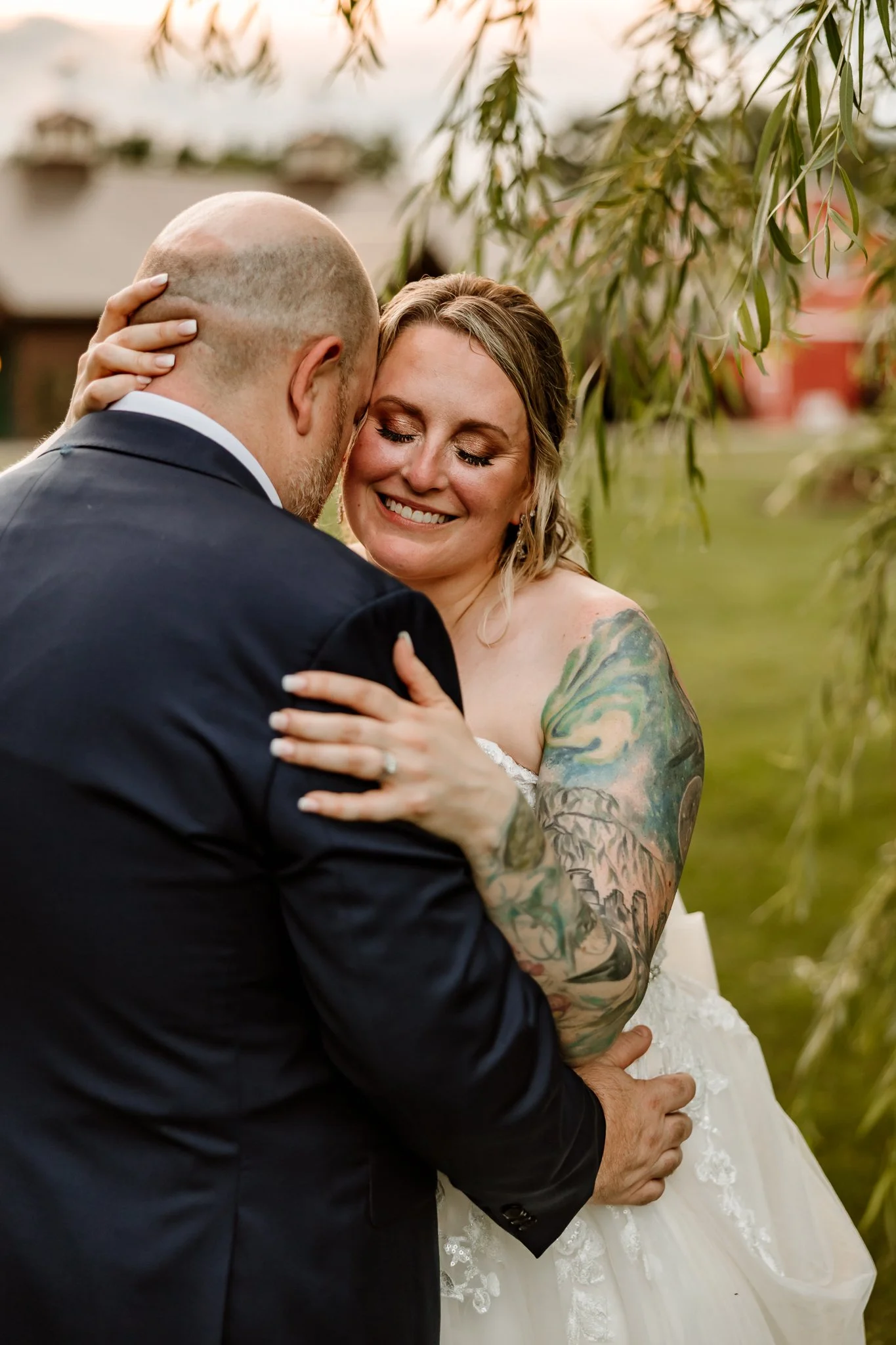 A bride and groom embrace outdoors, smiling happily, with one woman's visible tattooed arm, surrounded by greenery at sunset.