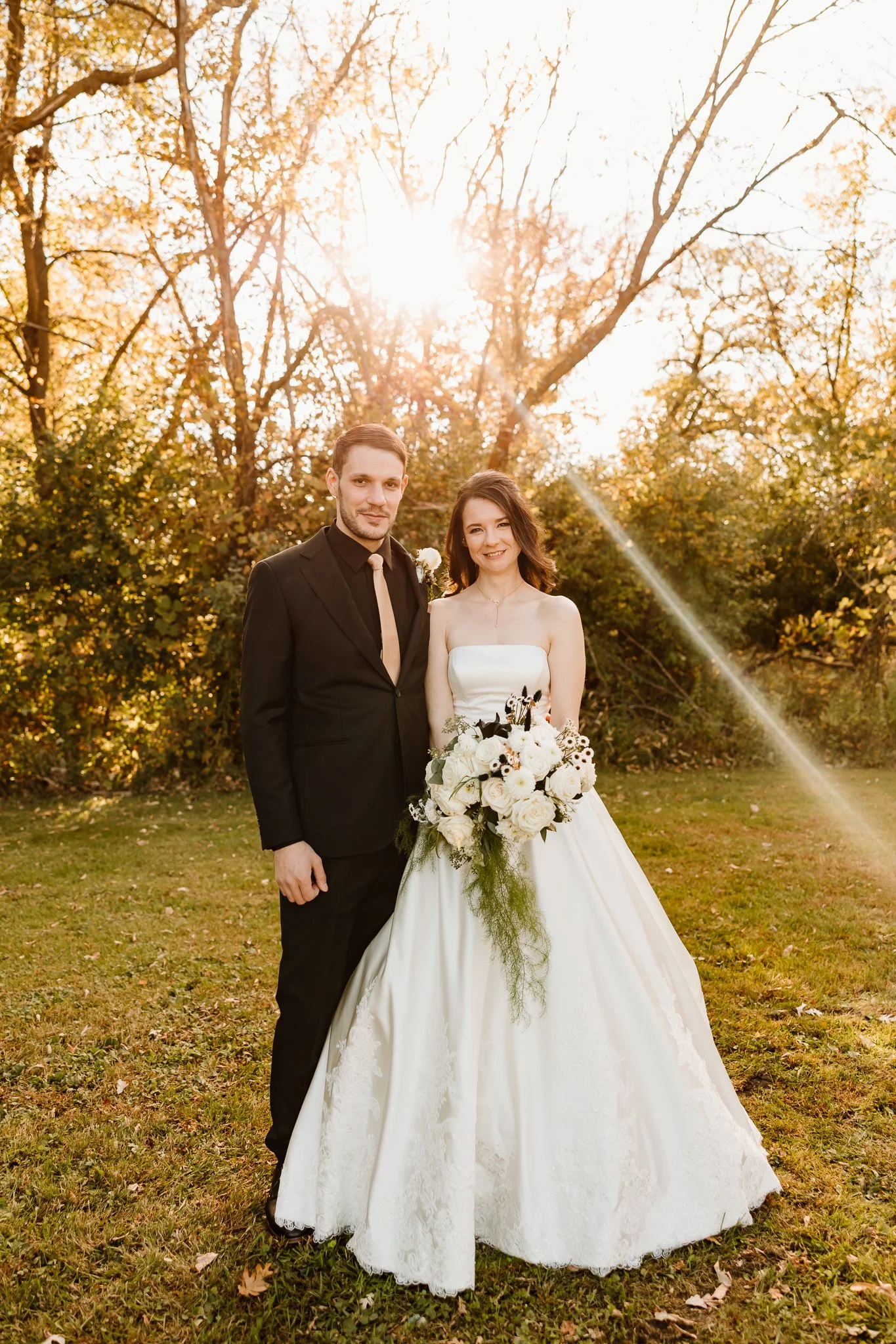 A bride and groom standing outdoors on a grassy area with trees in the background, during sunset. The bride is wearing a white strapless wedding gown and holding a bouquet of white flowers. The groom is dressed in a black suit with a light-colored ti