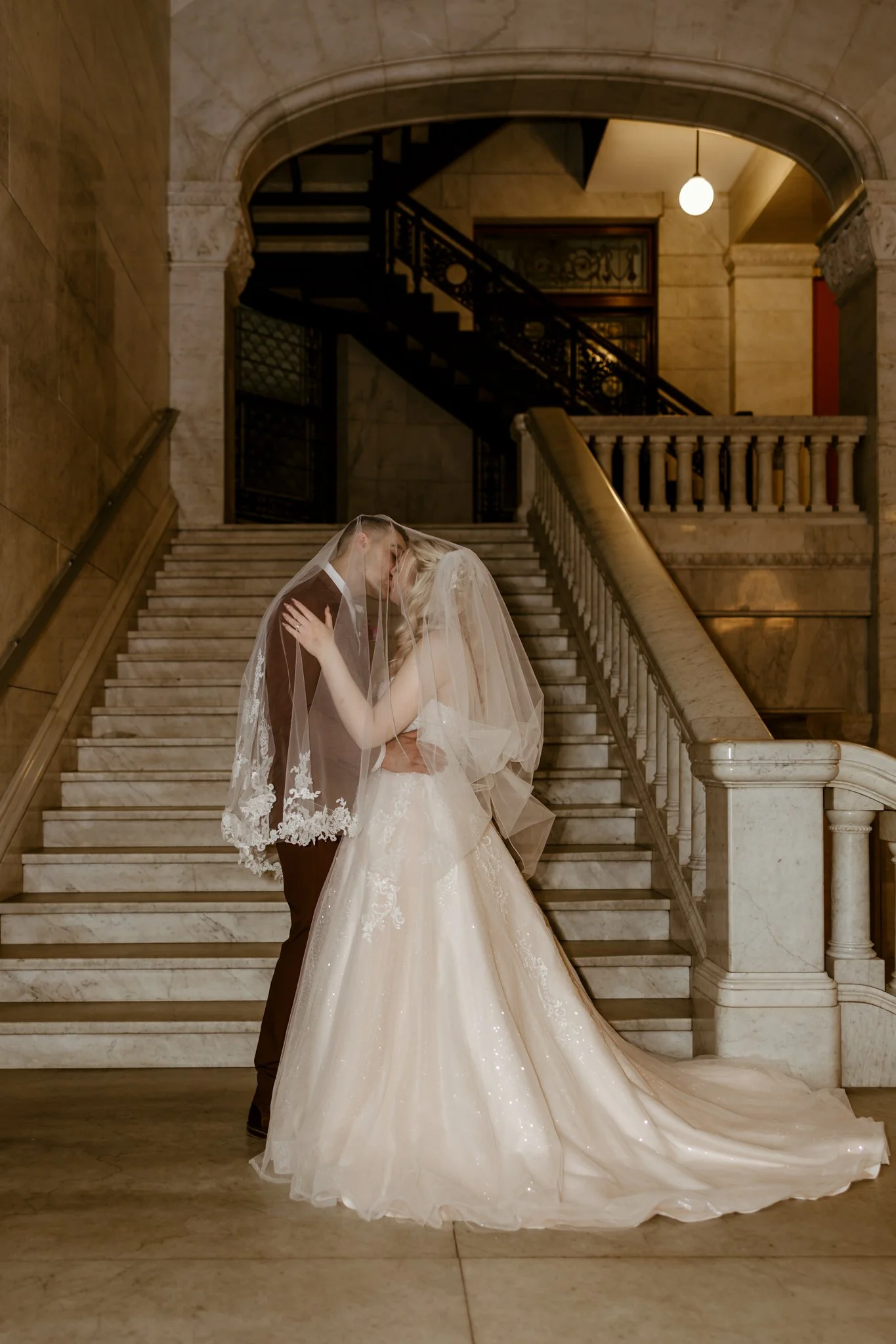 A bride and groom kissing on a grand staircase inside a building, with the bride wearing a white wedding gown and veil, and the groom in a suit.