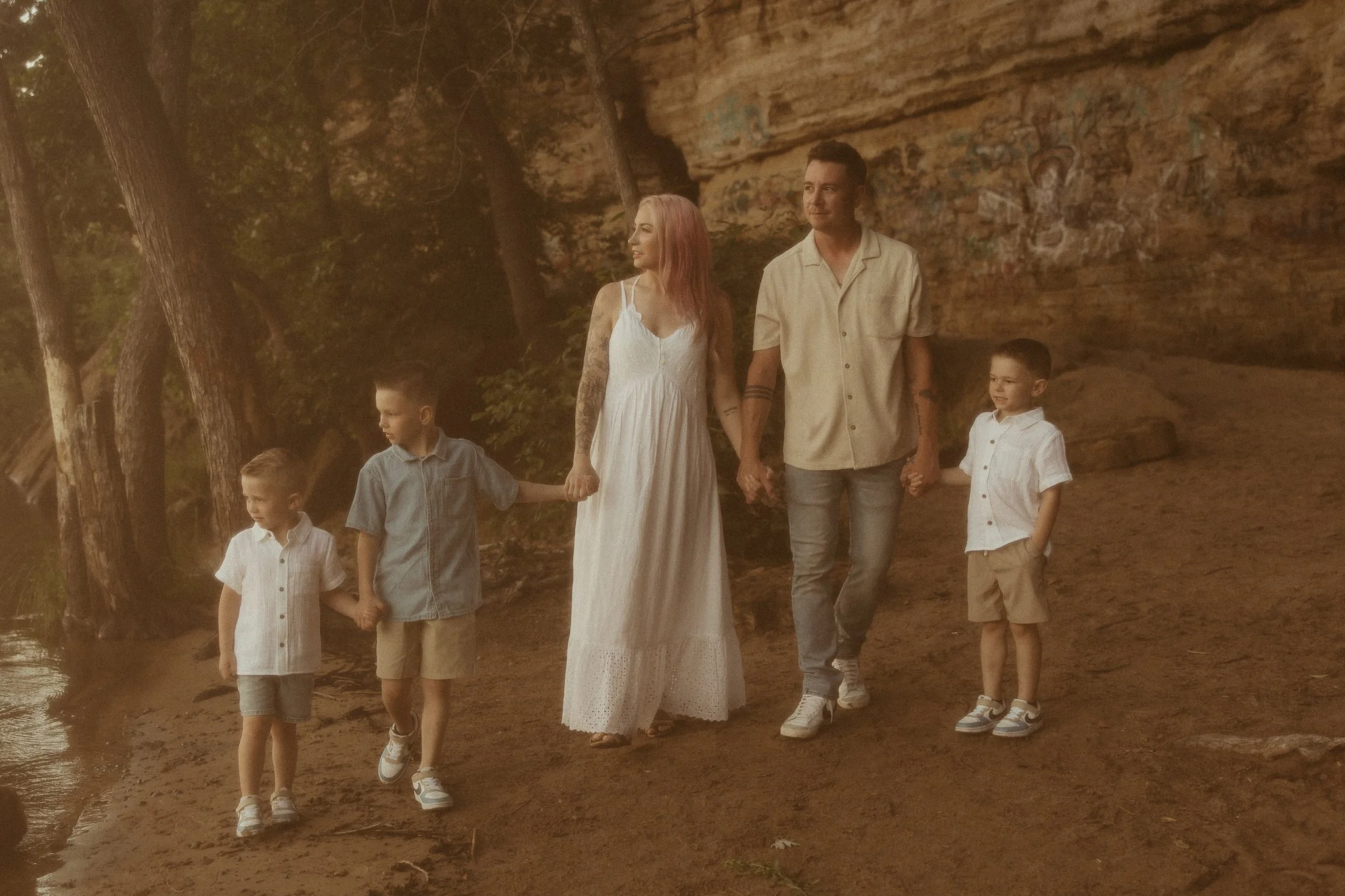 A family of five holding hands and walking along a dirt path near a body of water in a wooded area with rock formations in the background.