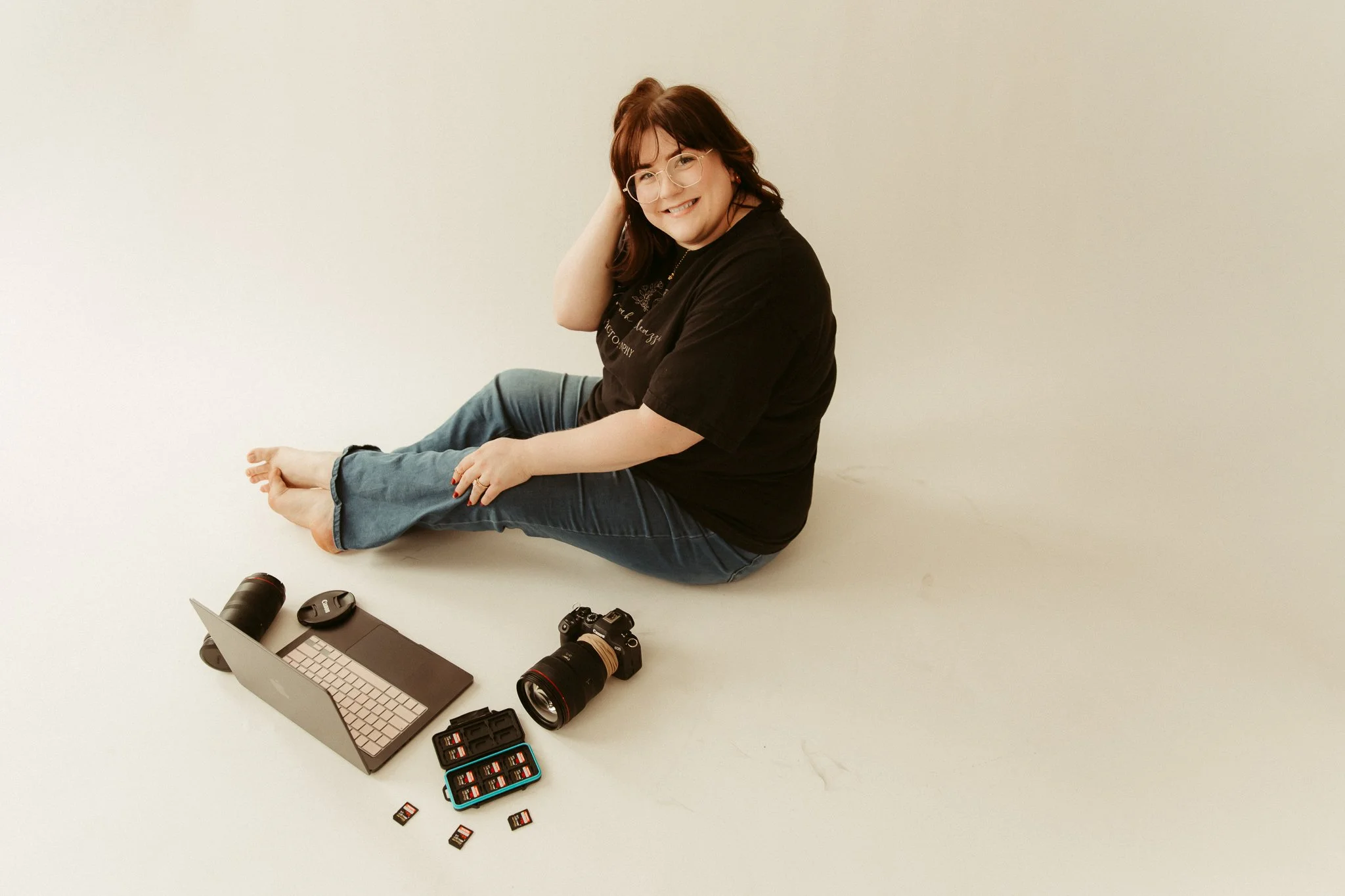 A woman with dark hair and glasses sitting on the floor, smiling. Cameras, a laptop, SD cards, and camera accessories are arranged around her on the floor.