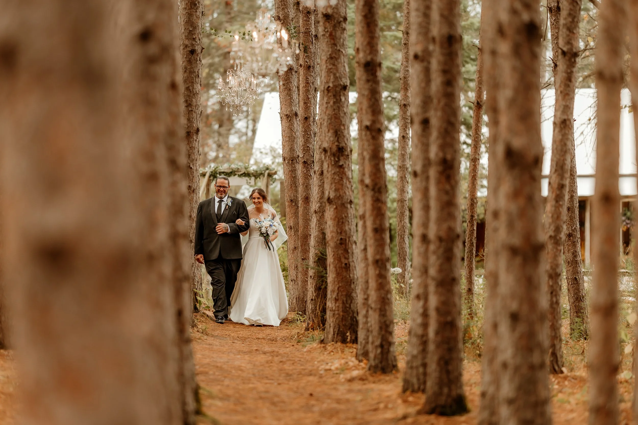Bride walking down a forest path with her father, holding a bouquet of white flowers, as chandeliers hang from the trees in an outdoor wedding setting. Bigfork mn wedding photography