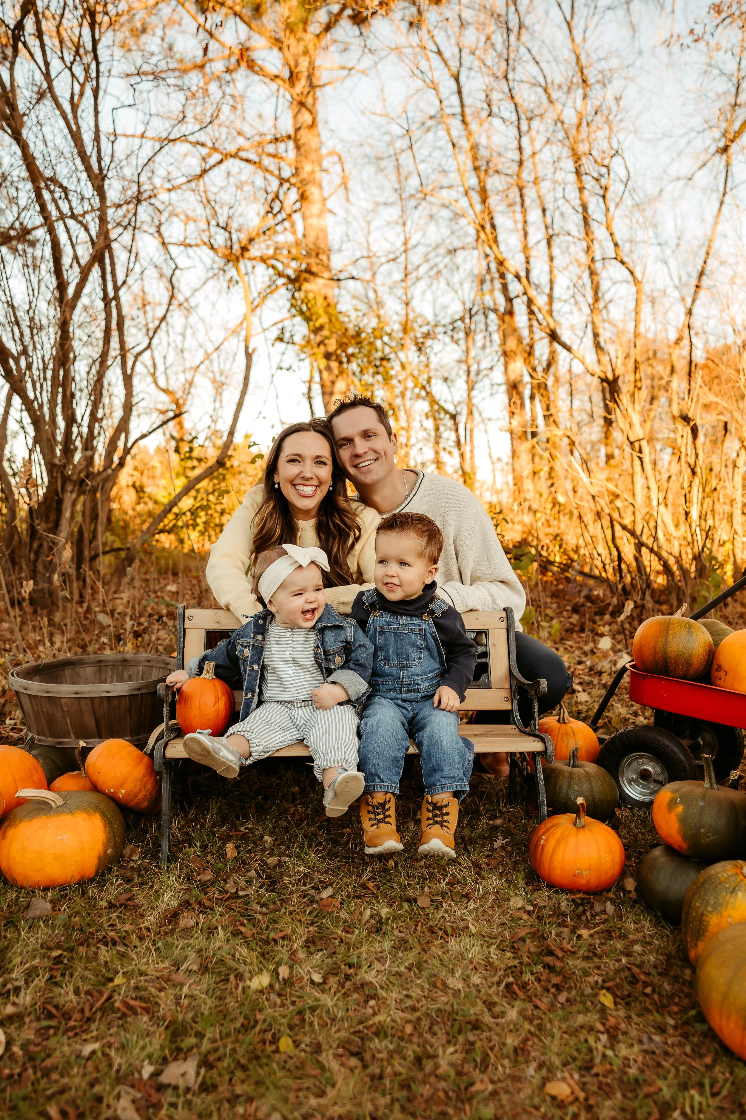 A happy family of four, including a mother, father, young girl, and young boy, sitting on a wooden bench surrounded by pumpkins in an outdoor fall setting, with leafless trees and a warm sunset in the background.