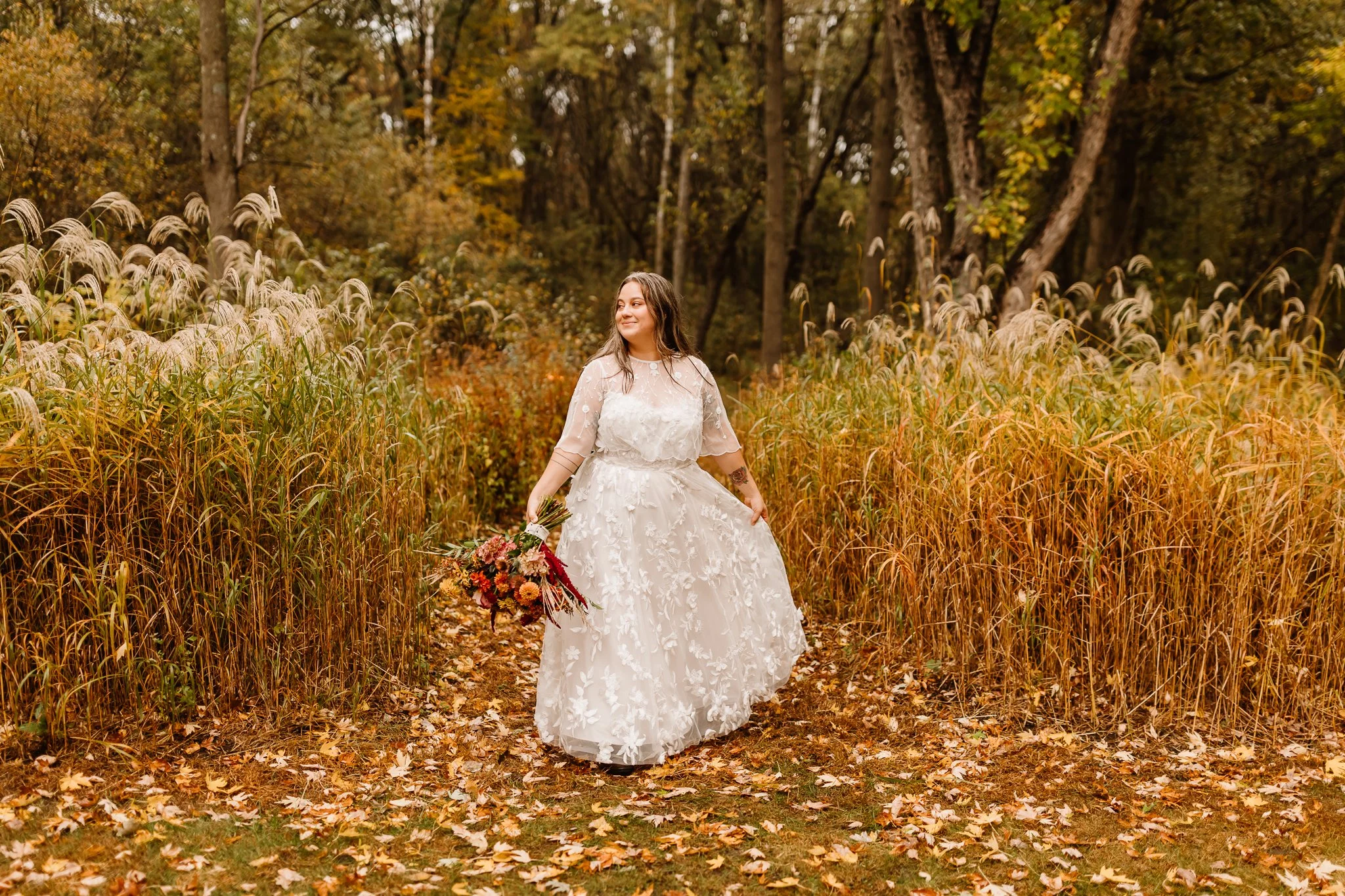 A woman in a white floral wedding dress holding a bouquet of flowers walking through a forest in autumn.
