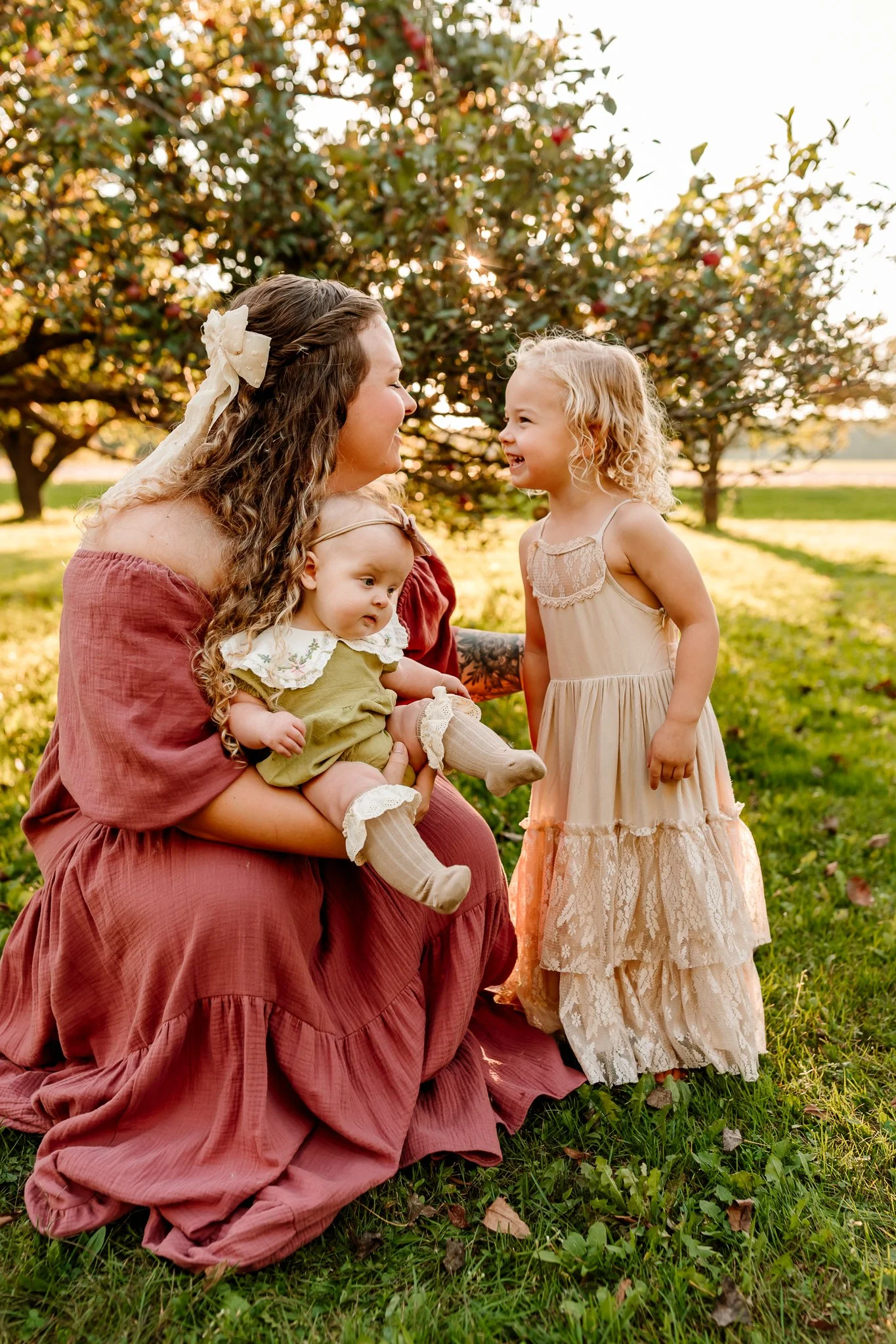 A woman with long curly hair and a large bow in her hair, dressed in a flowing pink dress, is sitting on the grass outdoors, holding a young child in green clothing. A little girl in a cream-colored lace dress stands in front of them, smiling and tal