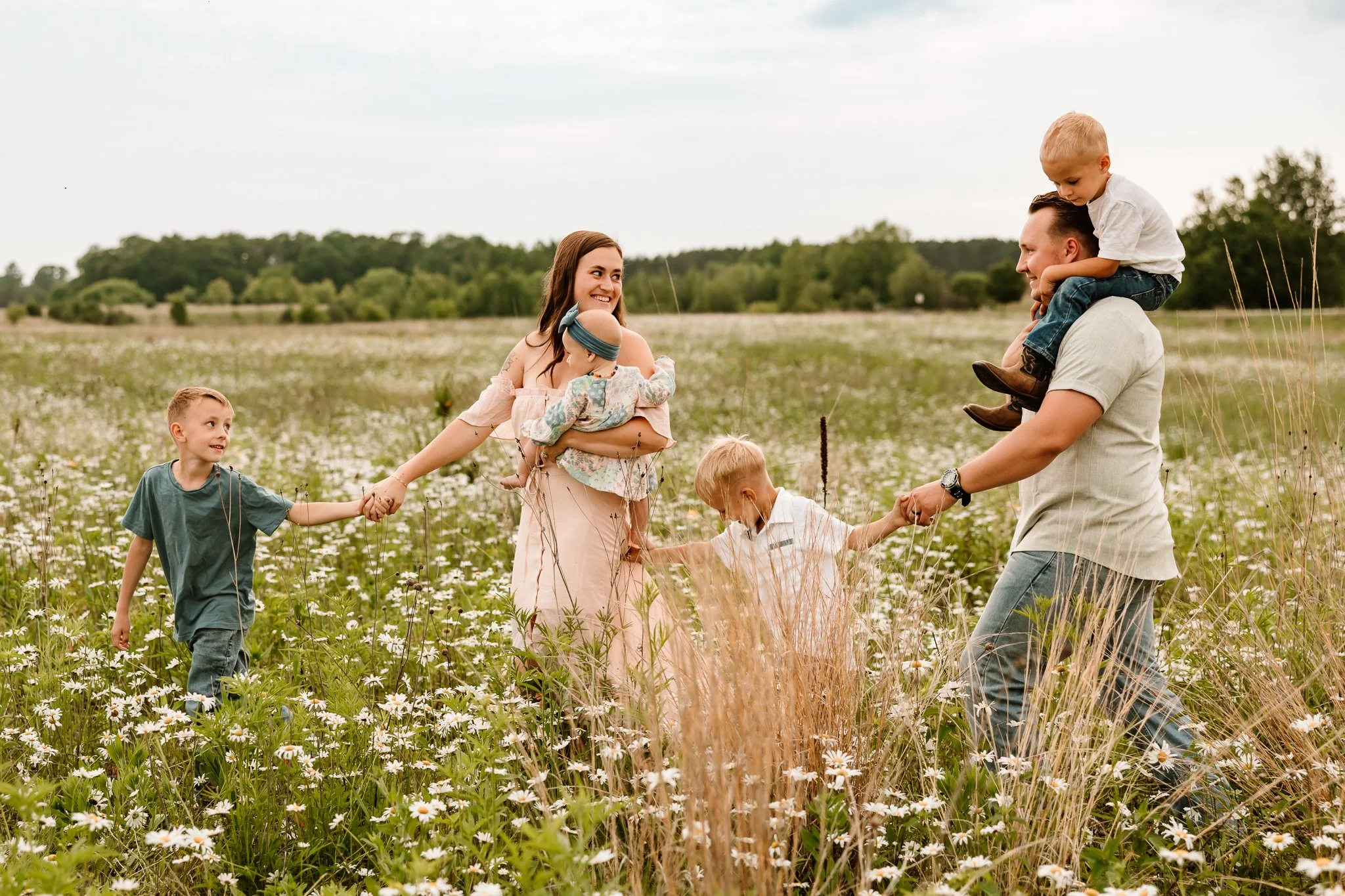 A family of six holding hands and playing in a field of wildflowers and tall grasses, with a backdrop of trees and sky.