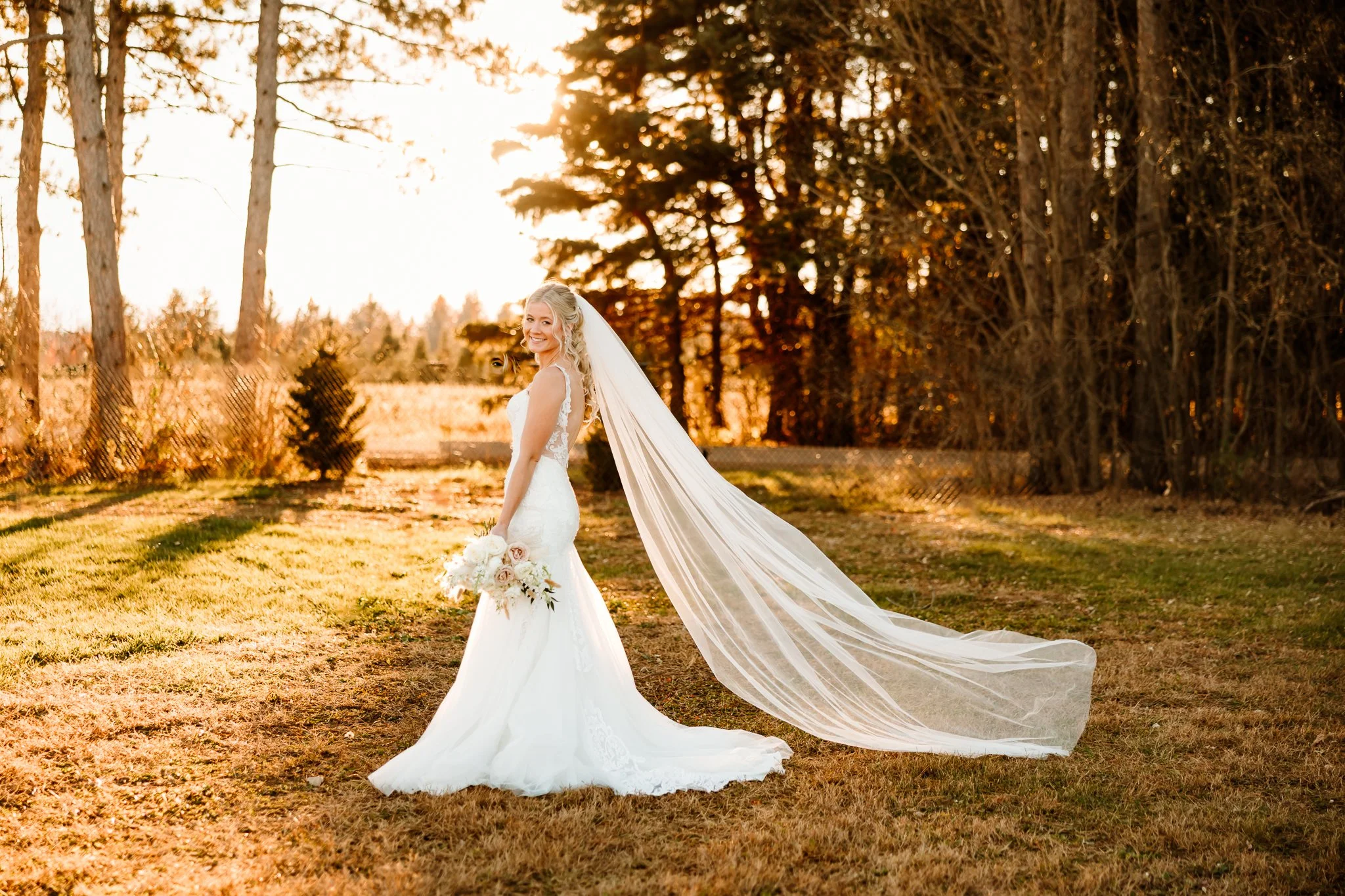 A smiling bride in a white wedding gown with a long veil, holding a bouquet, standing outdoors on grass with trees and sunlight in the background. Cambridge Minnesota 