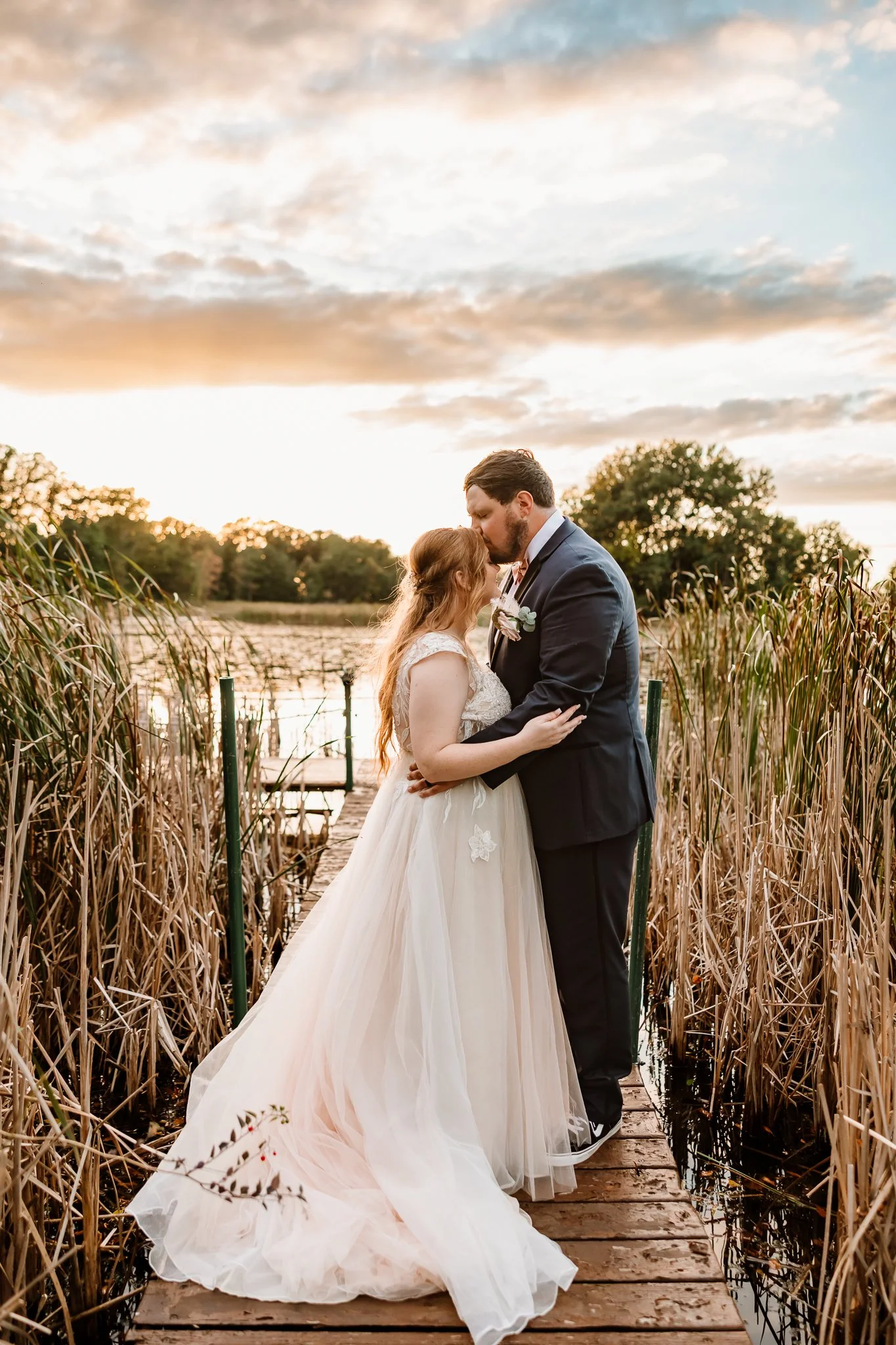 A bride and groom sharing a kiss on a wooden dock by a lake, surrounded by tall reeds and trees, during sunset.