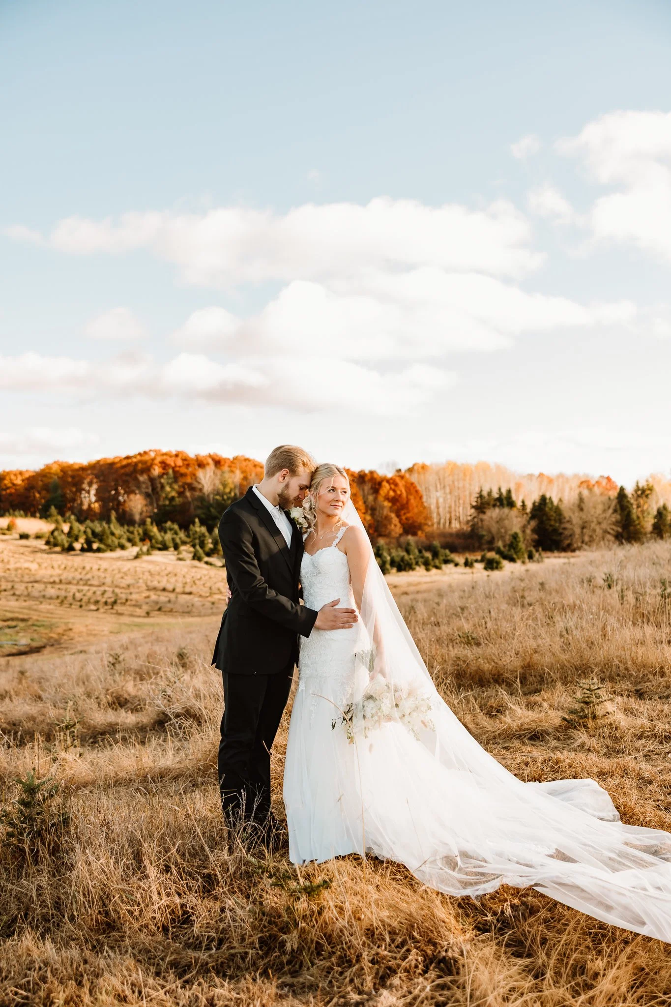 A bride and groom standing close together in a field during sunset, with trees in the background displaying fall colors.