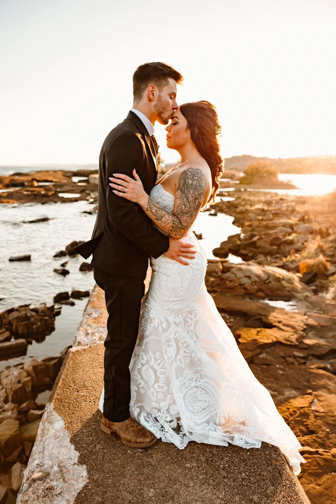 A bride and groom standing on a rocky shoreline at sunset, embracing and sharing a kiss.