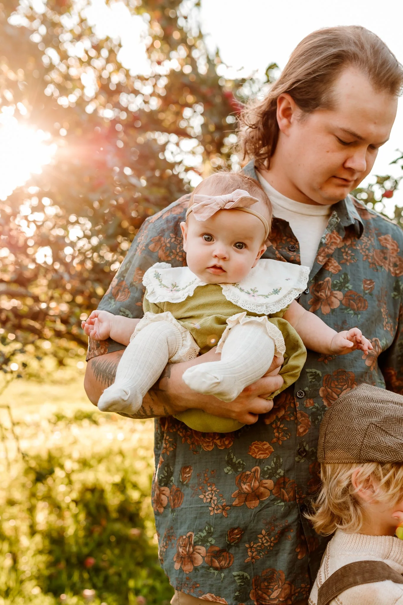 A man holding a baby girl outdoors during sunset, with another child partially visible at the bottom right, surrounded by trees and warm sunlight.