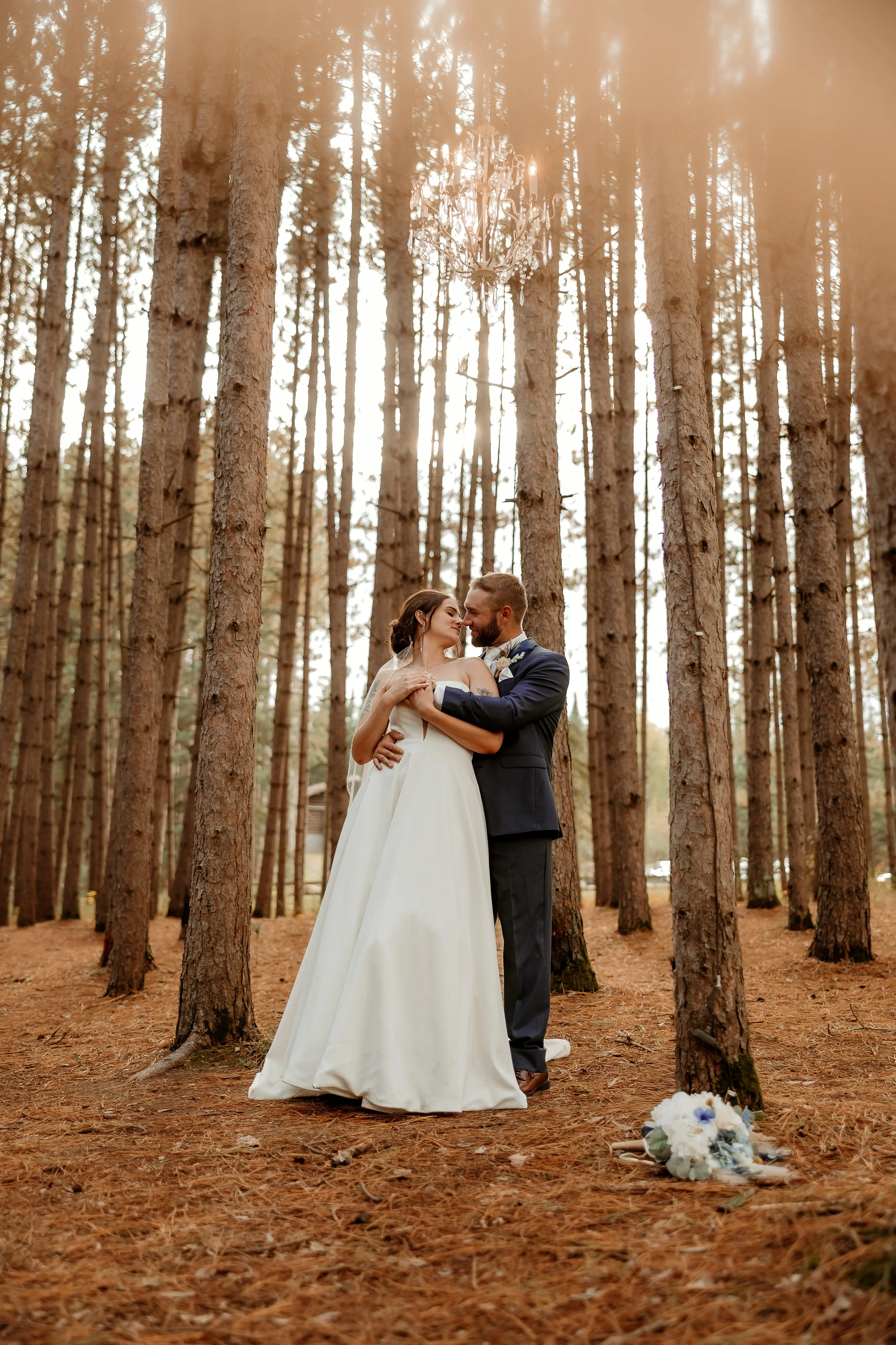 A bride and groom embrace in a forest with tall trees, a chandelier hanging from the trees, and a bouquet on the ground.