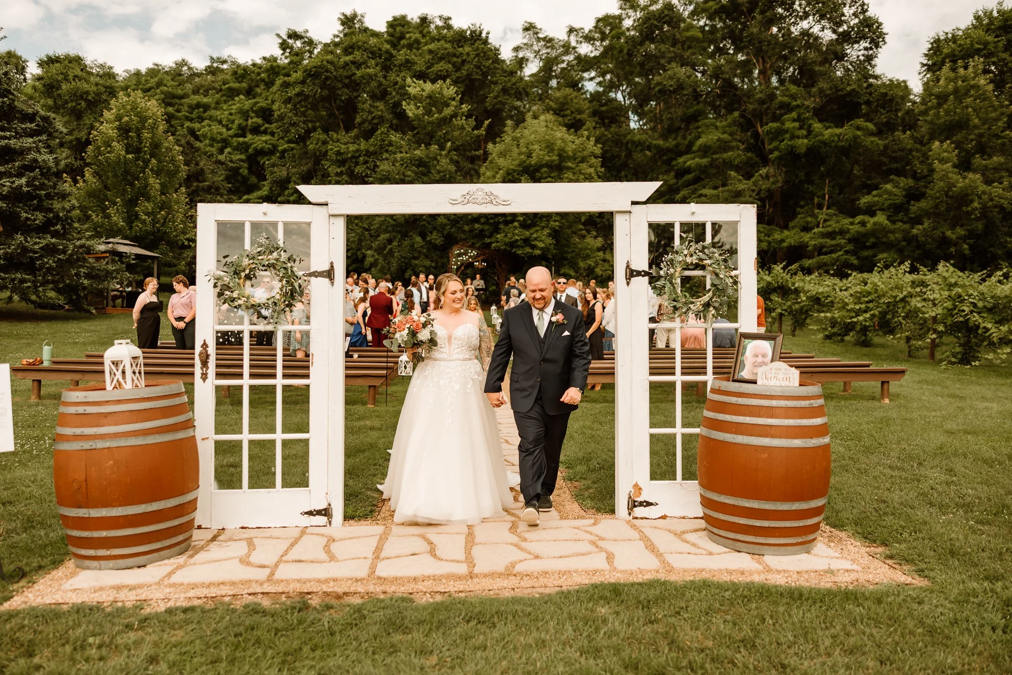 Bride and groom walking through a white wooden gate with glass windows and decorative wreaths at an outdoor wedding ceremony. Guests are in the background, seated on benches, amidst greenery and trees.