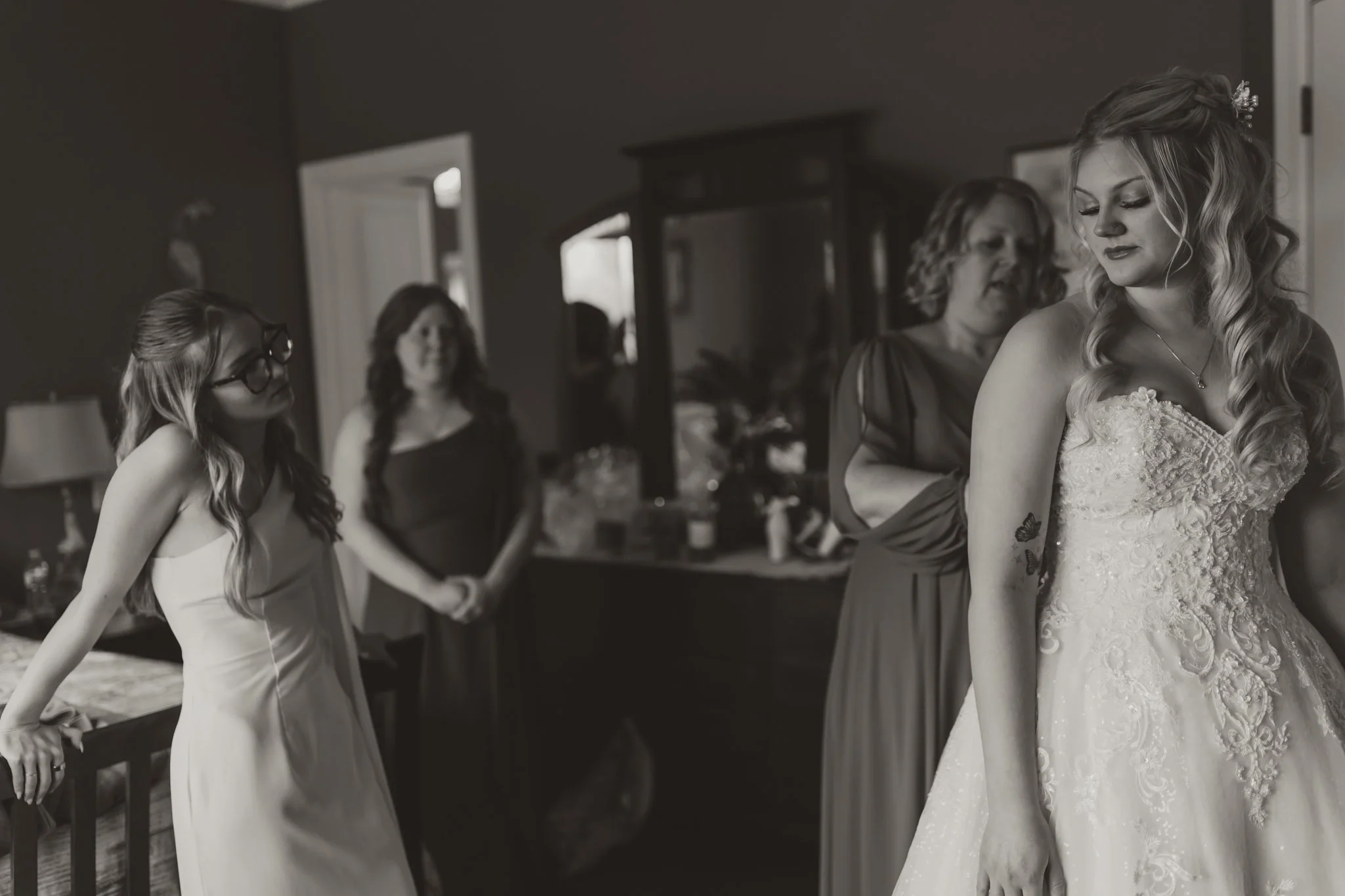 A bride in a wedding dress with lace details getting ready with her bridesmaids and mother in a bedroom.