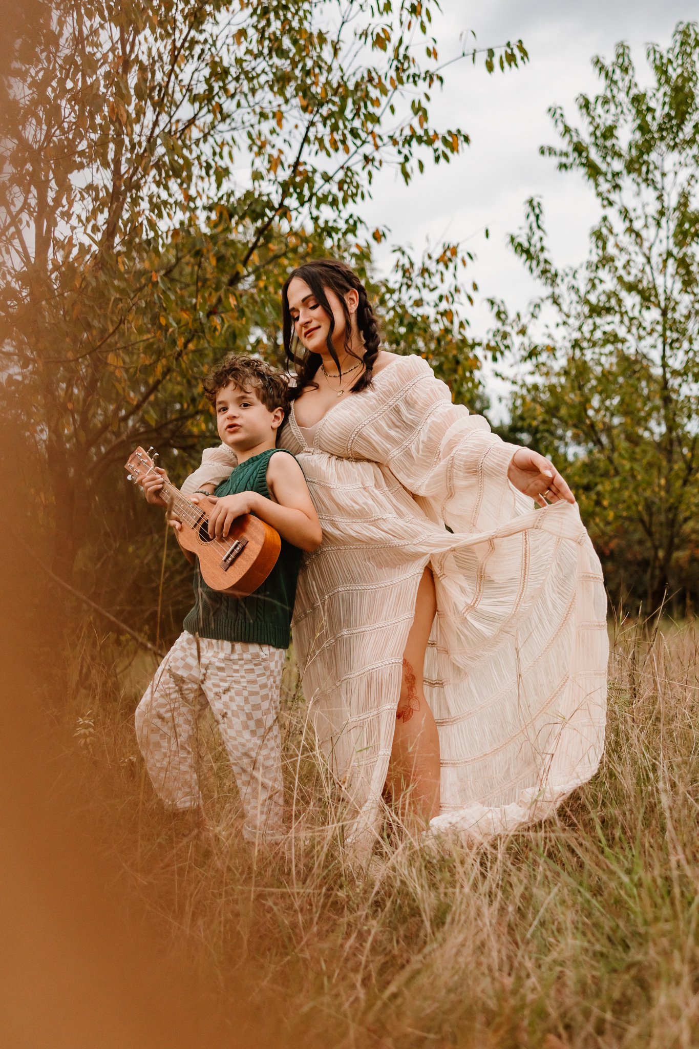 A woman in a flowing cream dress standing outdoors in a grassy field, with a young boy holding a ukulele standing beside her, both surrounded by trees.