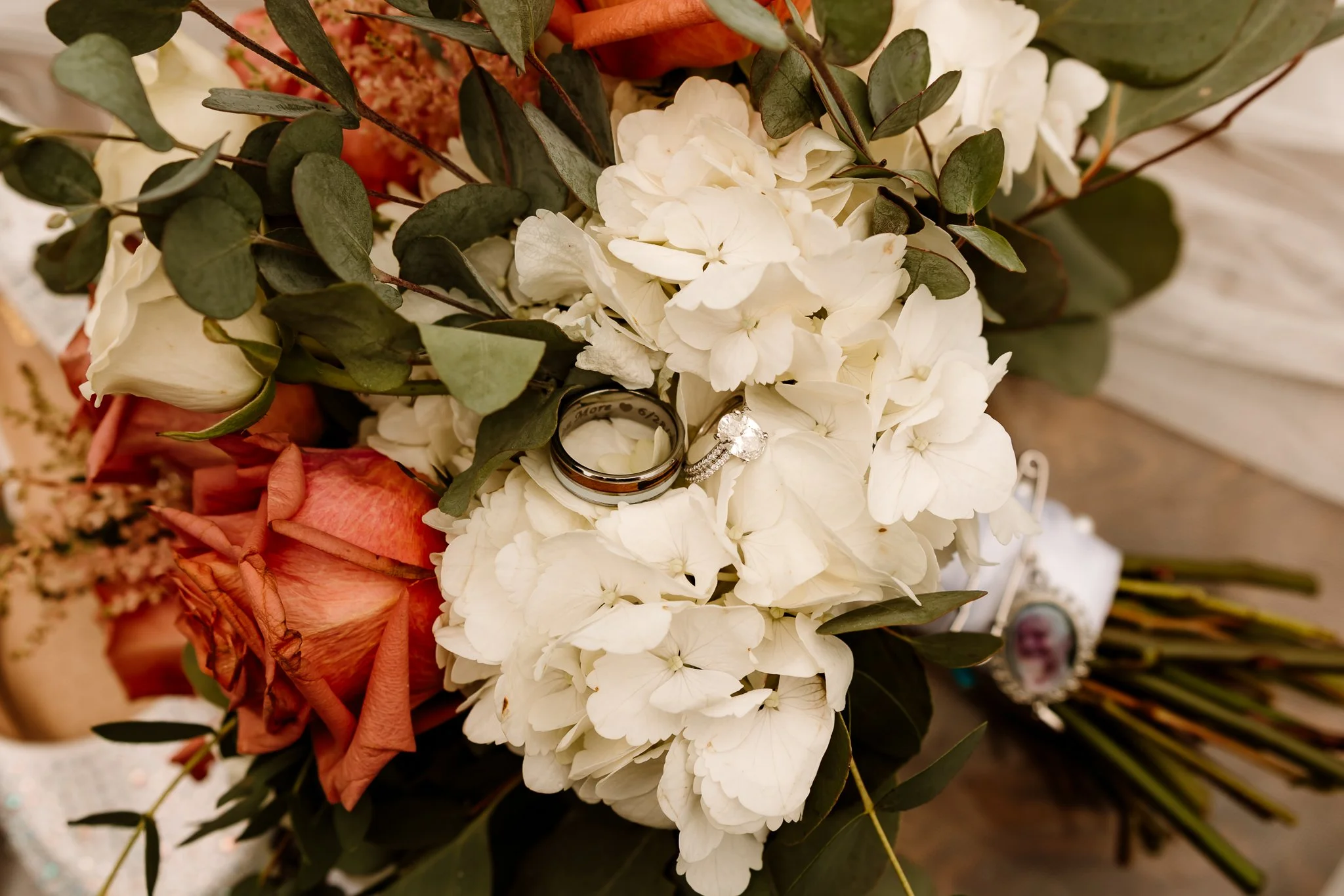 Wedding bouquet with white hydrangeas, green leaves, and peach-colored roses, featuring a wedding band and engagement ring placed on the white hydrangeas.