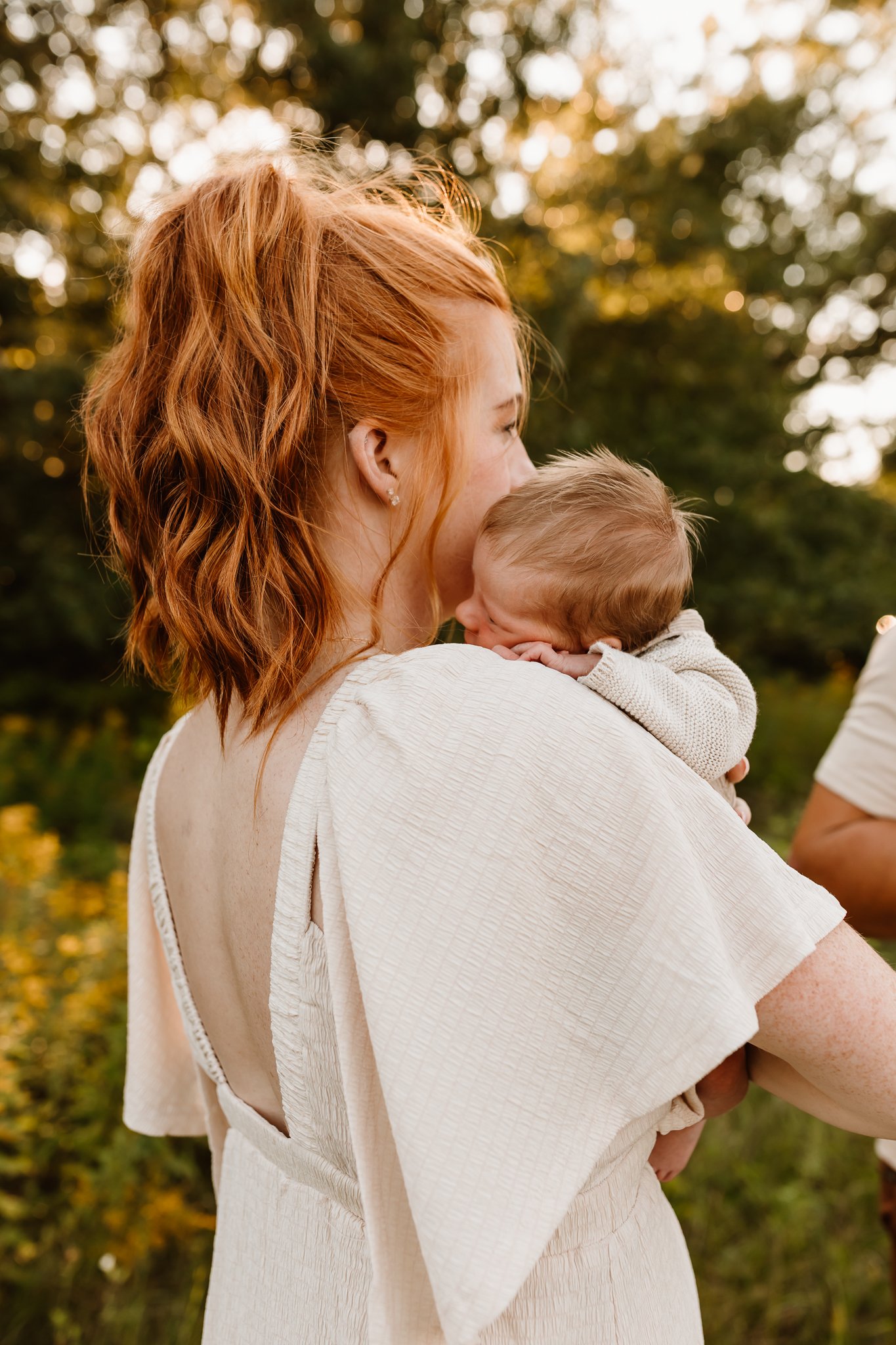 A woman with red hair holds a baby close to her shoulder outdoors during golden hour. She is wearing a light-colored dress with an open back, and the baby is resting face-down on her shoulder. The background features trees with autumn leaves.