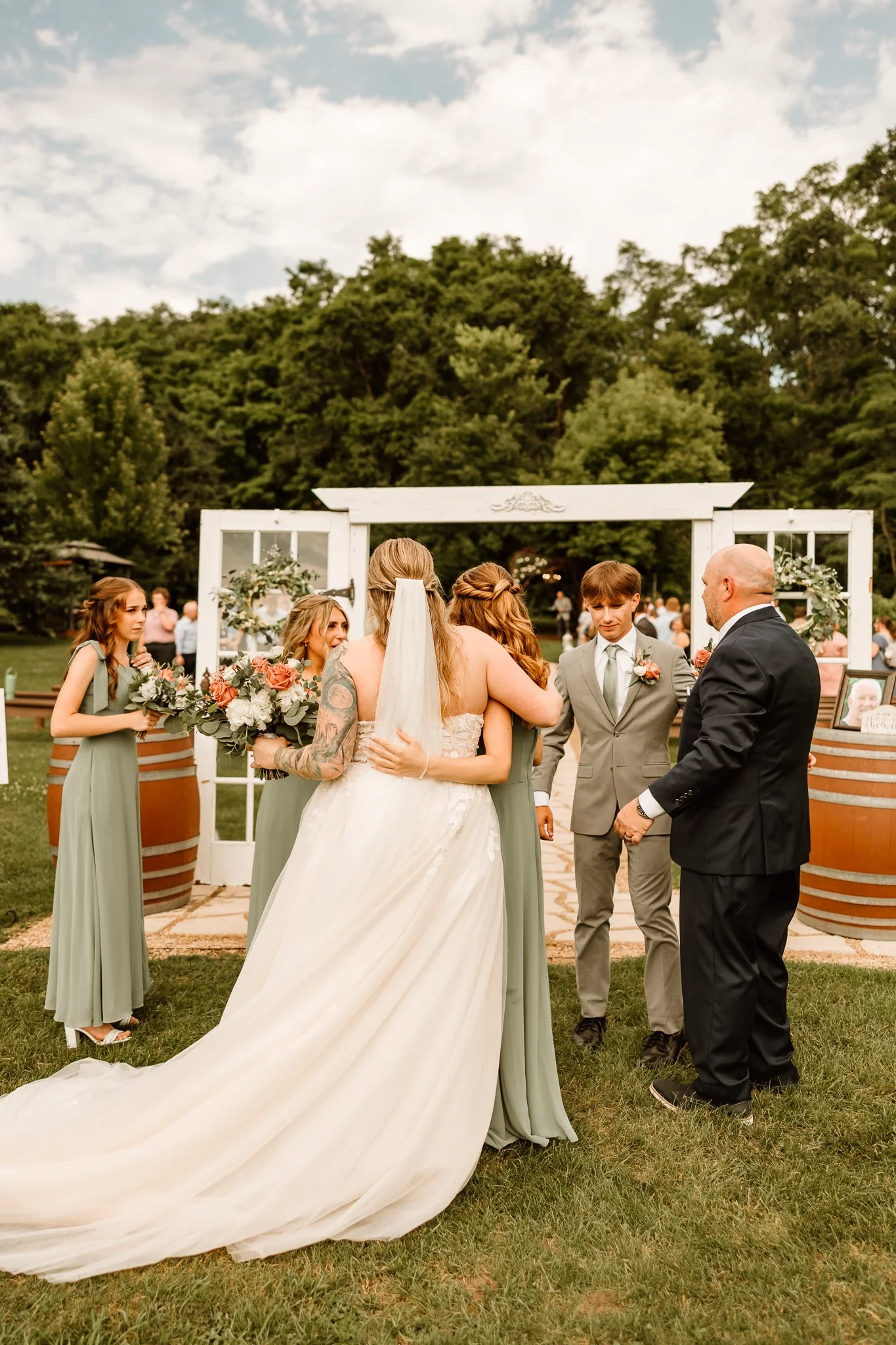 A bride in a cream wedding gown hugging a bridesmaid in a sage green dress at an outdoor wedding ceremony. Groomsmen and additional bridesmaids are nearby, with a white floral arch and greenery in the background.