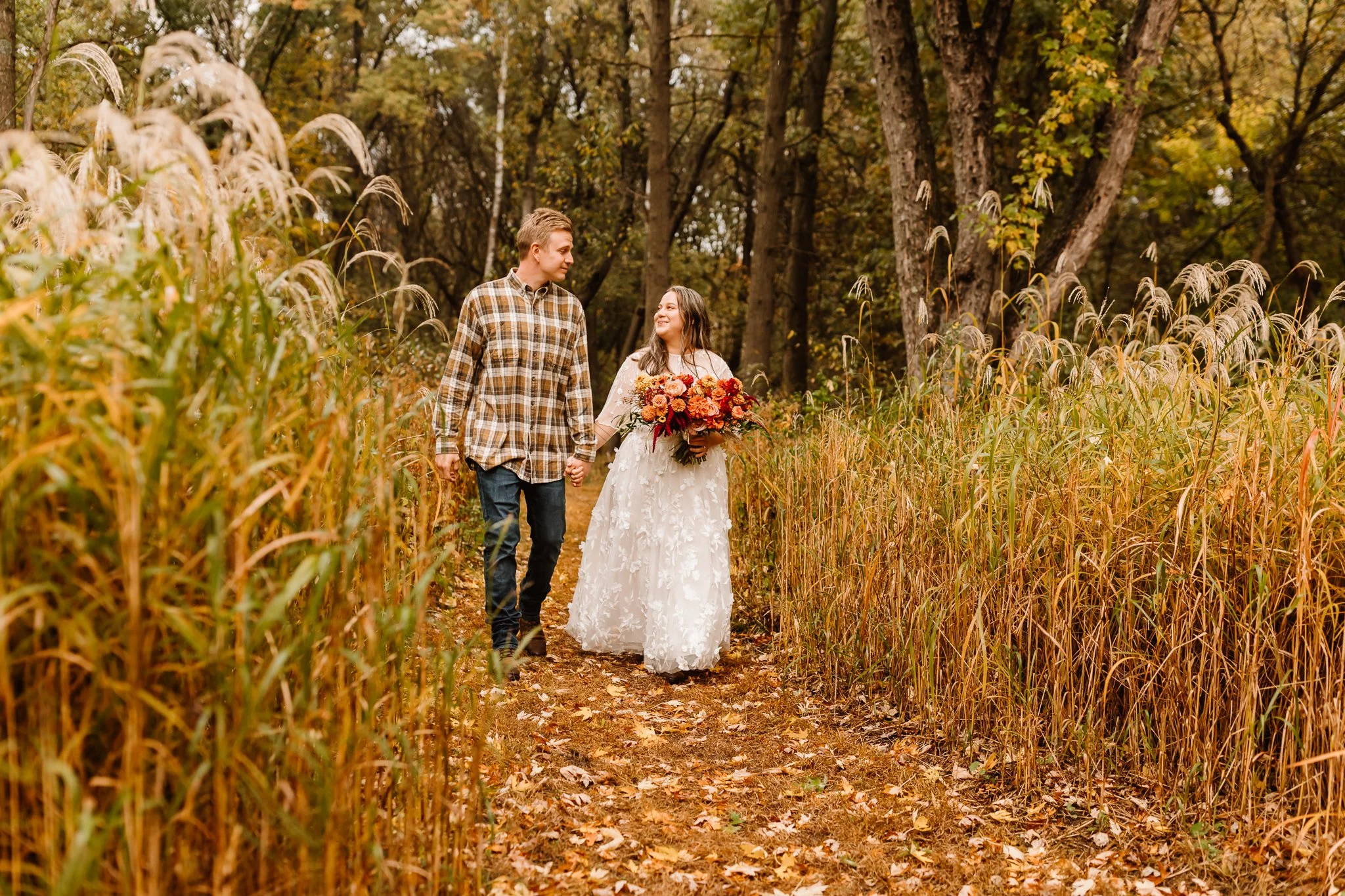A newlywed couple walking hand-in-hand through a fall forest trail, with tall grass and trees with colorful autumn leaves.