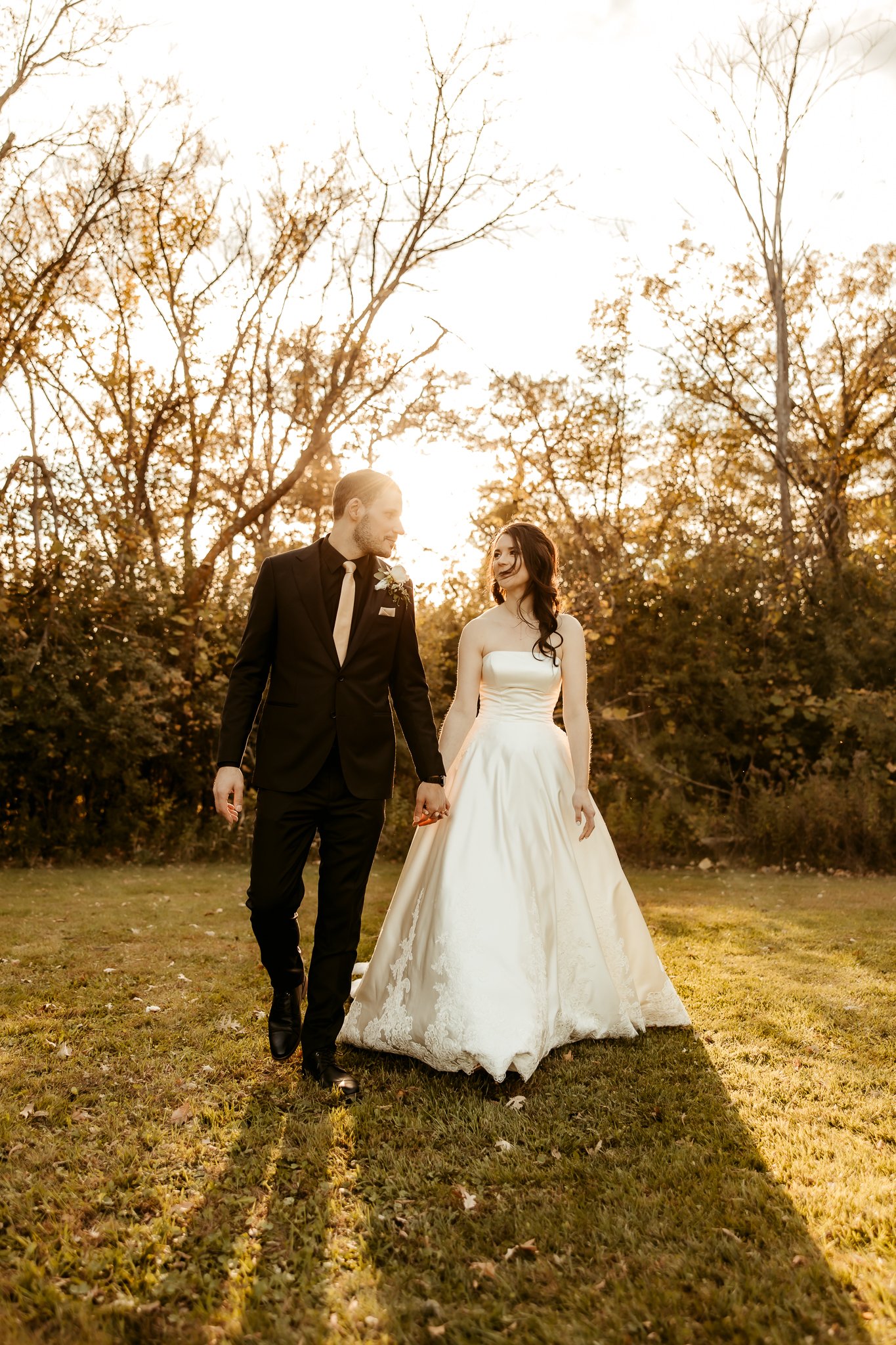 A bride and groom walking hand in hand outdoors during sunset, surrounded by trees.