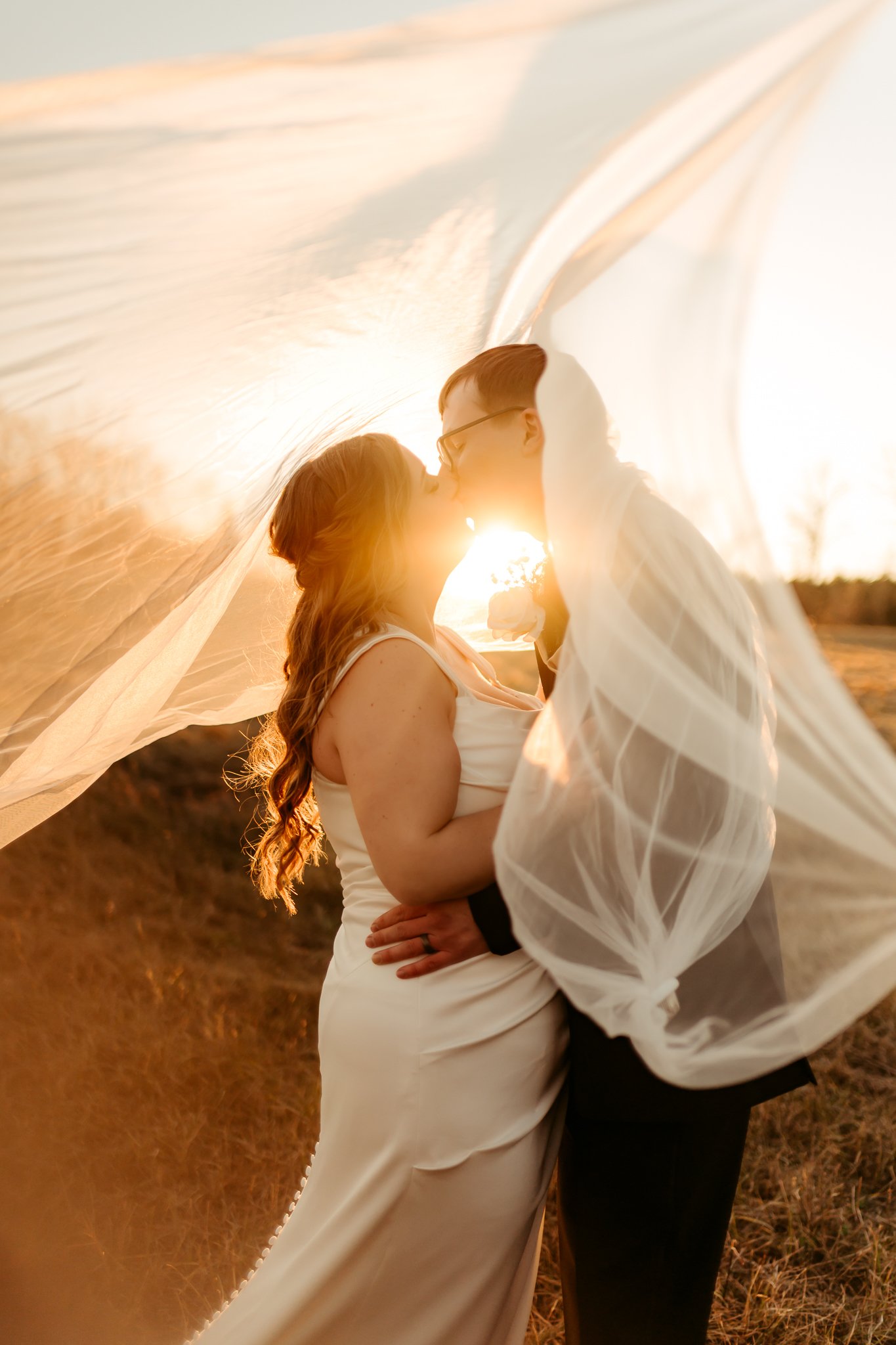 A couple sharing a kiss during sunset, with the bride in a white dress and the groom in a suit, standing outdoors under flowing fabric.