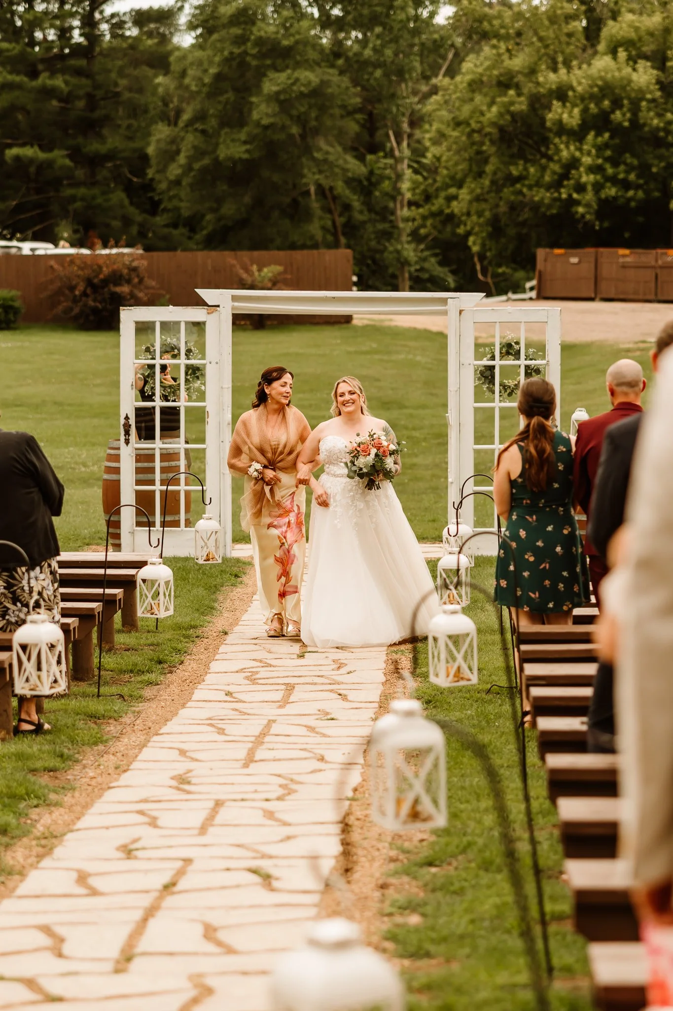 A bride walking down the outdoor aisle with her mother at her wedding ceremony, surrounded by guests seated on either side, decorated with white lanterns and flowers, against a backdrop of green trees.