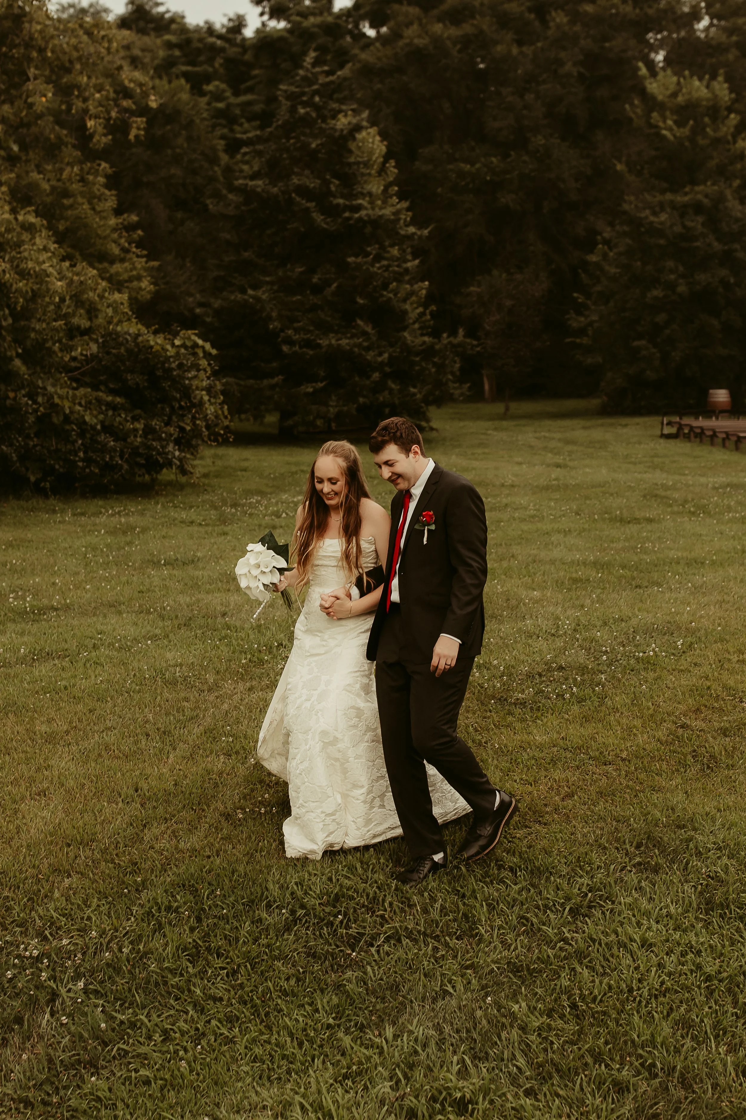 A bride and groom walking on grass during their wedding, holding hands and smiling with trees in the background.