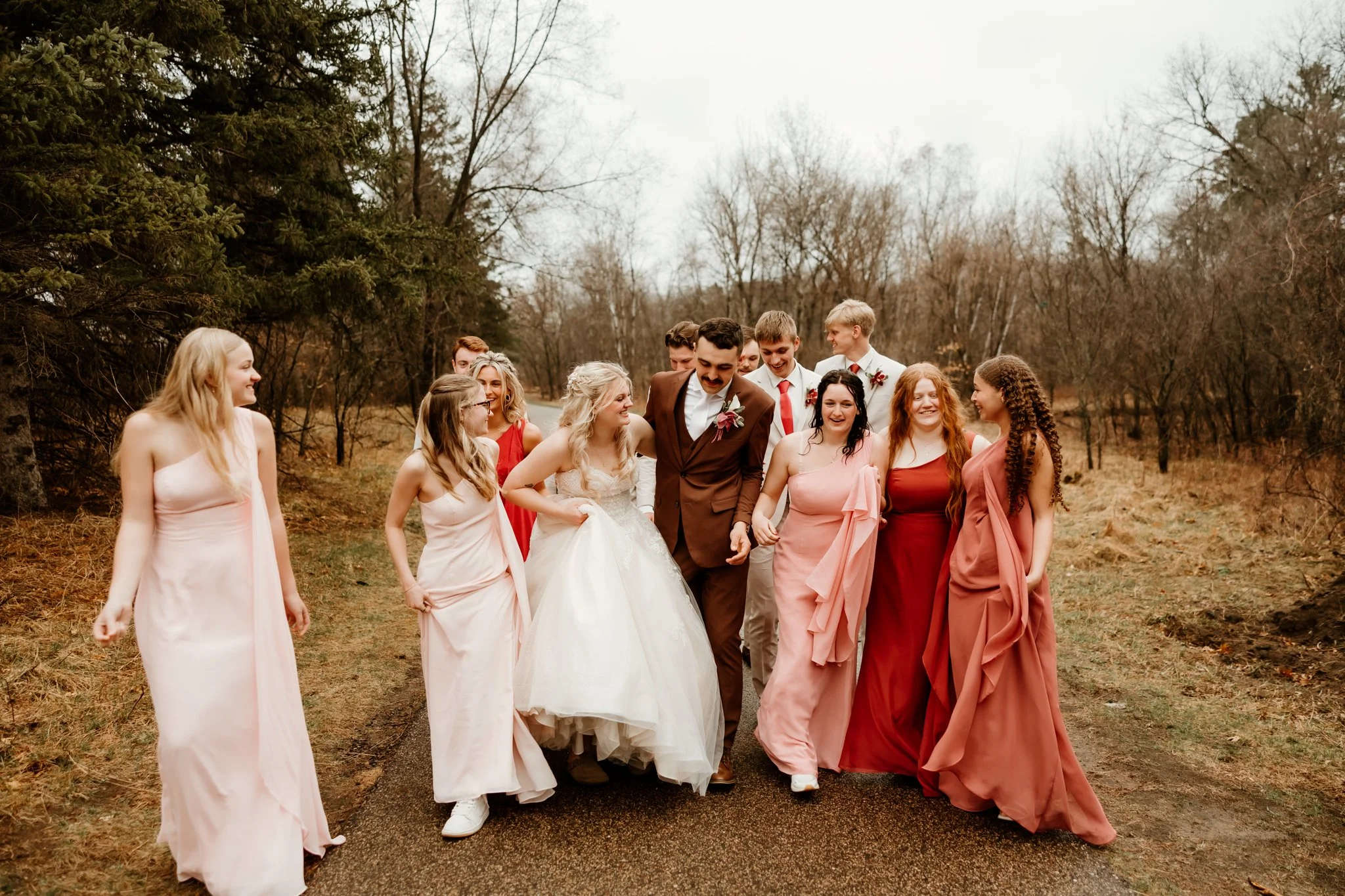 Group of people dressed in formal attire walking outdoors on a trail during autumn or winter. The group includes women in pastel and red dresses, and men in suits. The setting is a wooded area with leafless trees and overcast sky.